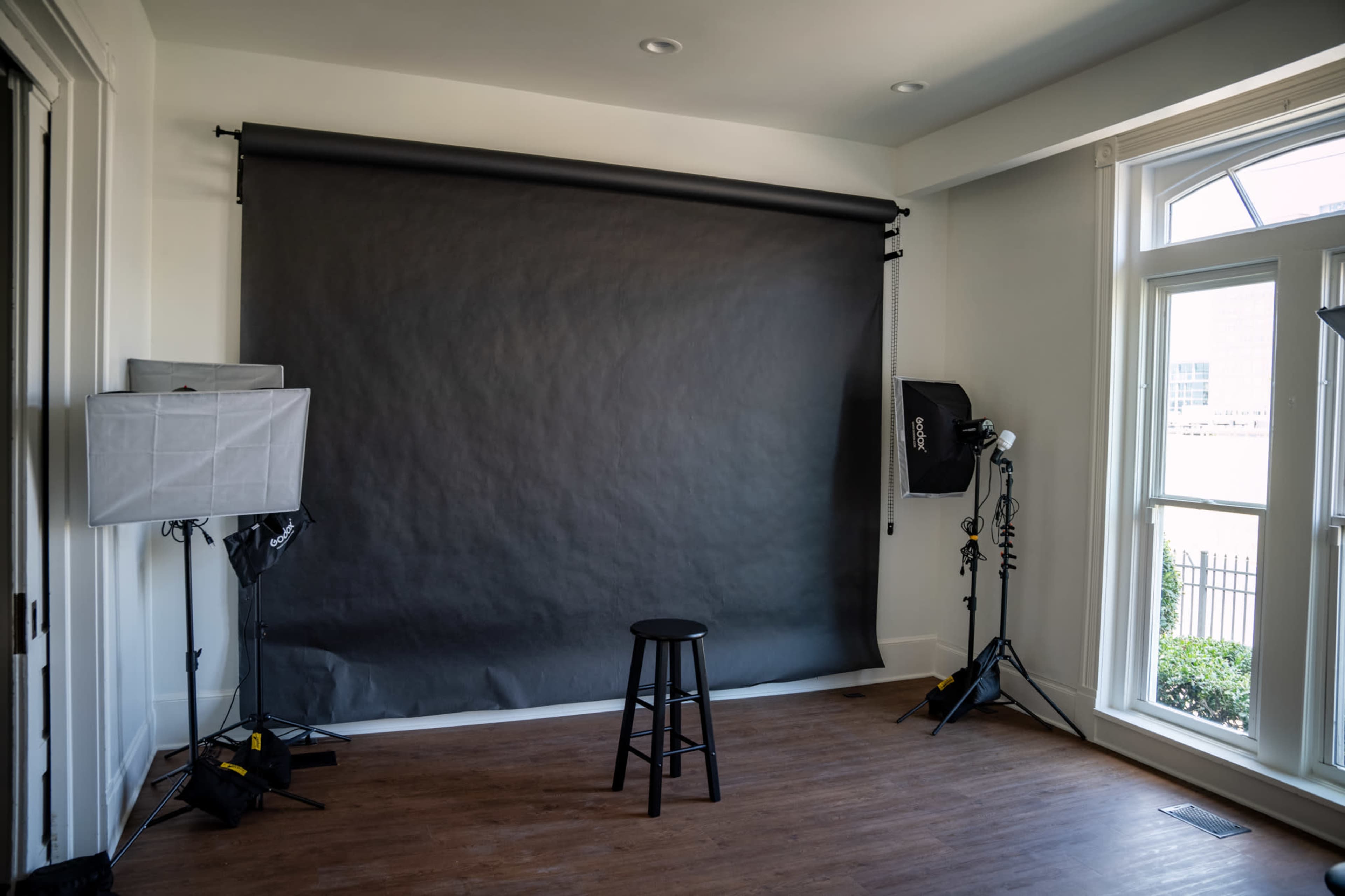 The image shows a photography studio with a dark backdrop, a black stool at the center, and lighting equipment positioned around the room.