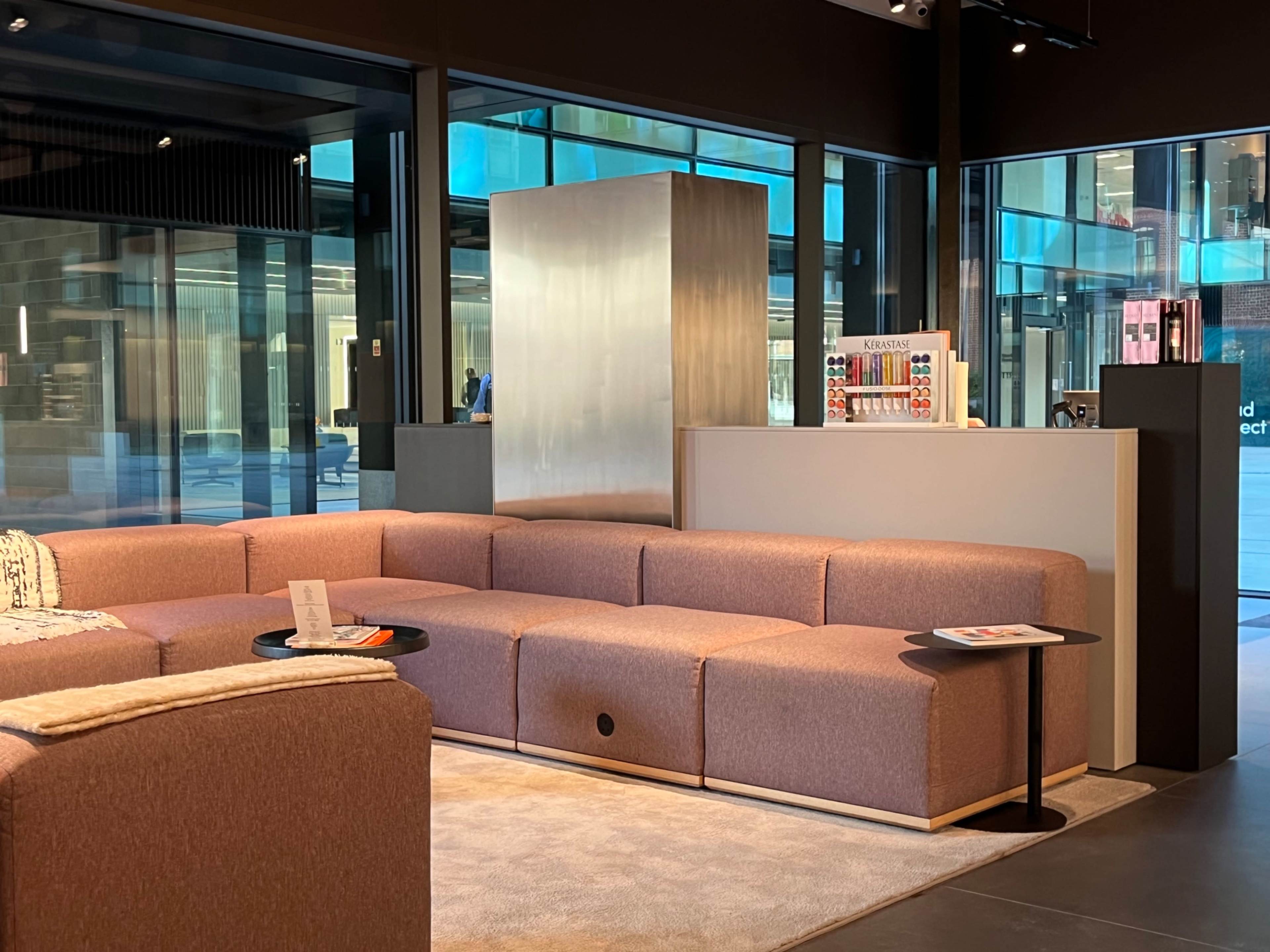 The image shows a modern lounge area with a large pink sectional sofa, a small black side table, and a sleek metallic structure in the background.
