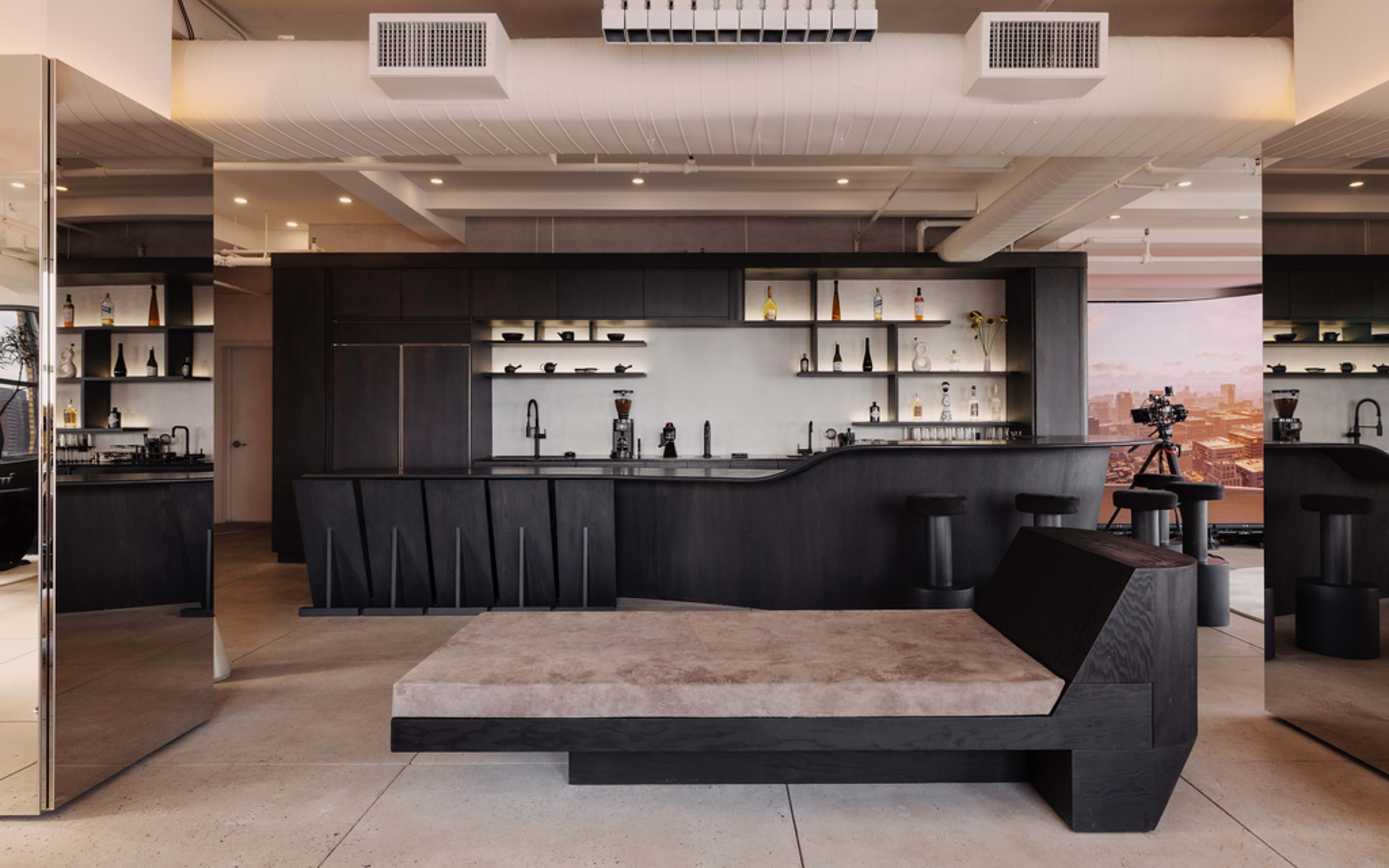 The image shows a modern, minimalist kitchen with dark cabinetry, a light counter space, and a sleek, low seating area in front of it.