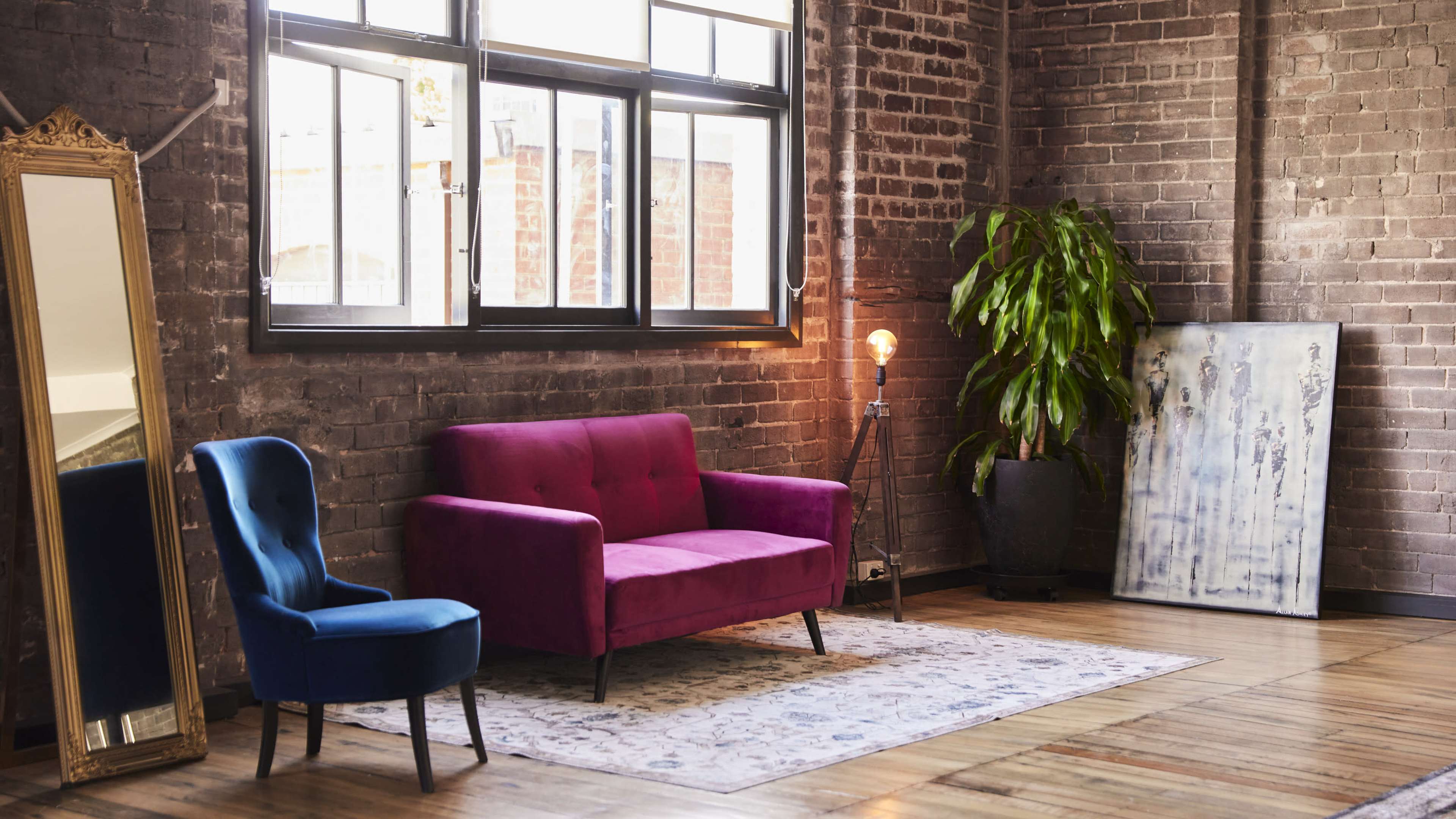 A pink sofa and a blue chair are arranged on a patterned rug in a softly lit room with exposed brick walls and large windows.