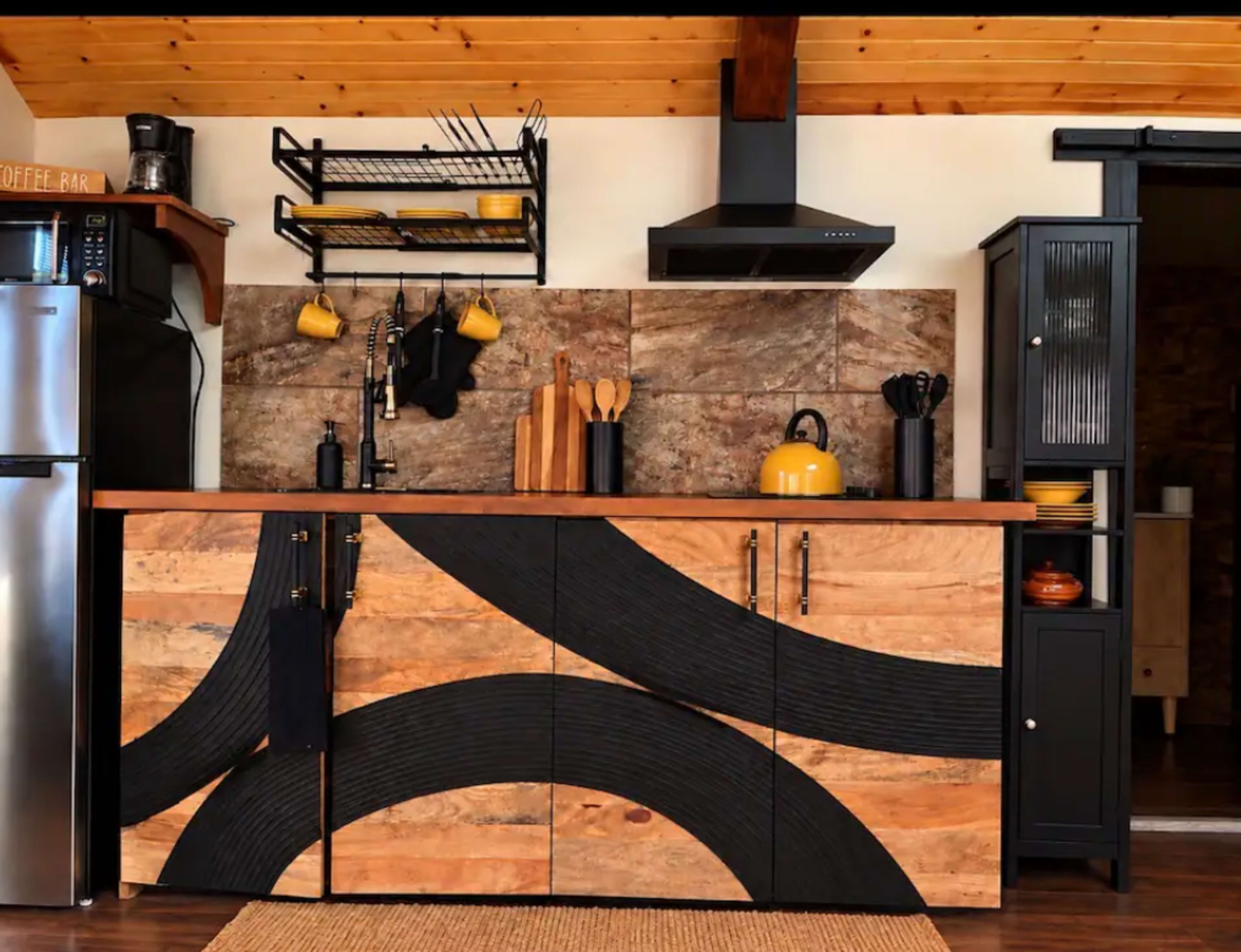 The image shows a modern kitchen with a wood and black cabinetry design, featuring open shelving, stainless steel appliances, and a range hood above the stove.