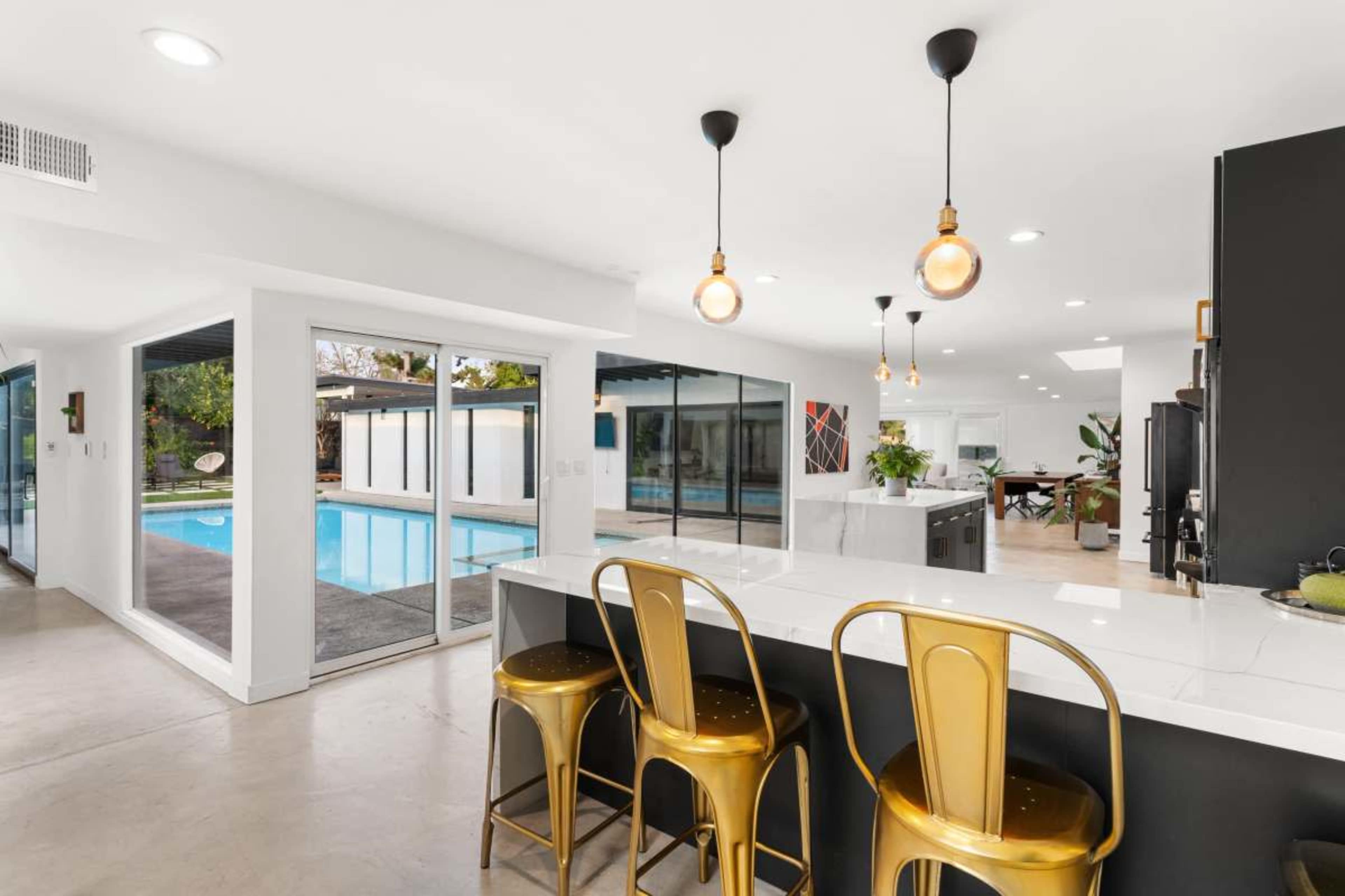The image shows a modern kitchen area with three gold bar stools overlooking a swimming pool through large glass doors.
