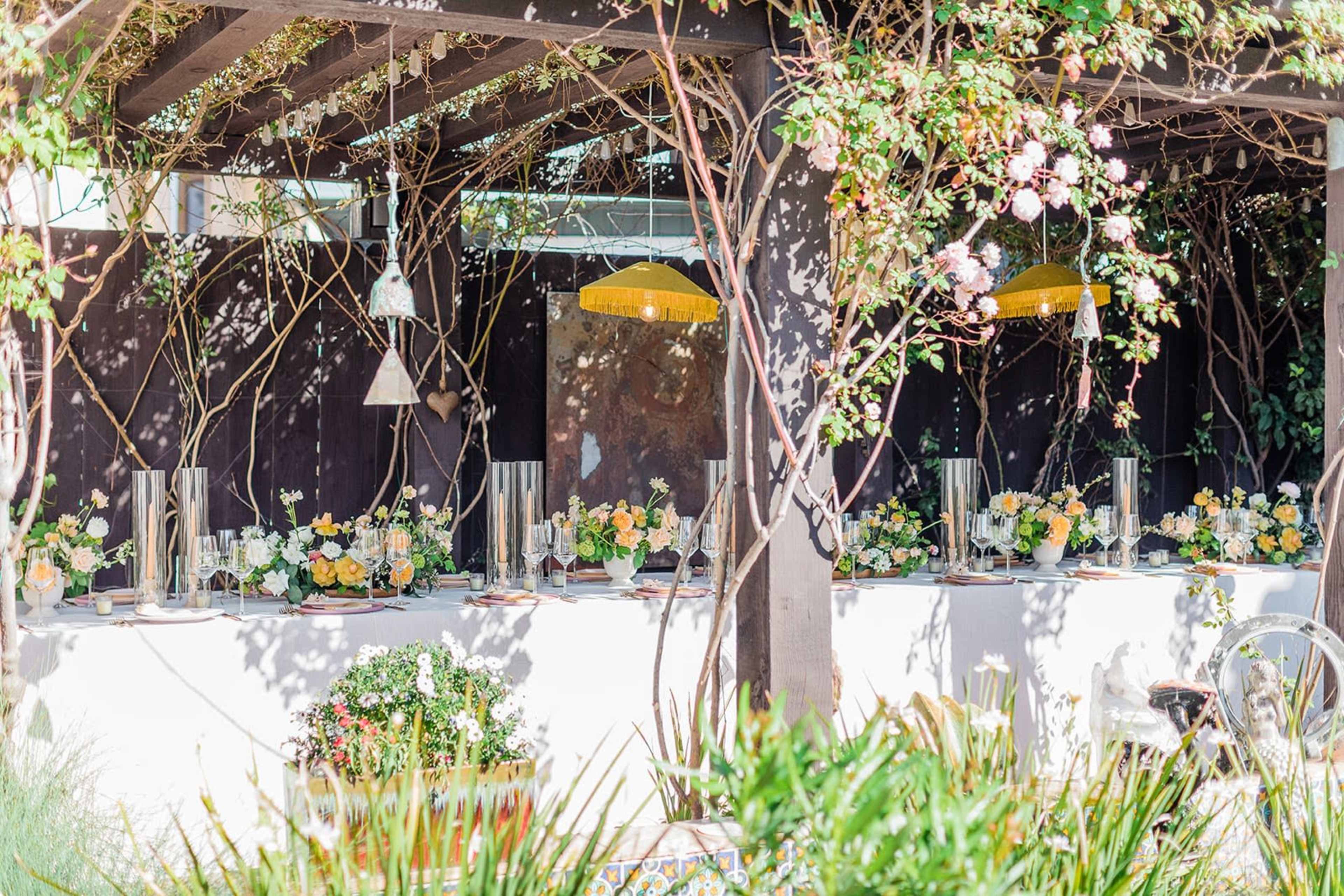 A decorated outdoor dining area features a long table set with floral arrangements and glassware under a pergola adorned with vines and hanging fixtures.