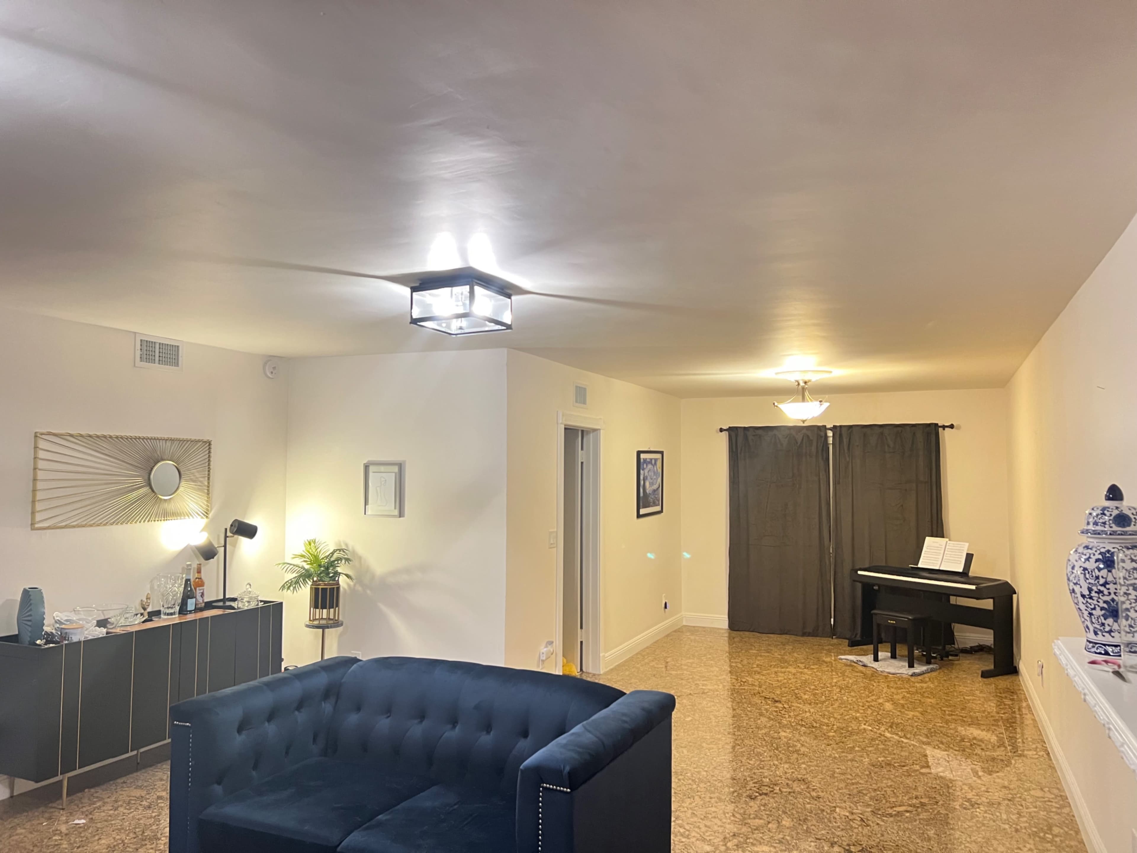 A spacious living room with a dark blue couch, a grand piano near a pair of dark curtains, and a modern light fixture on the ceiling.