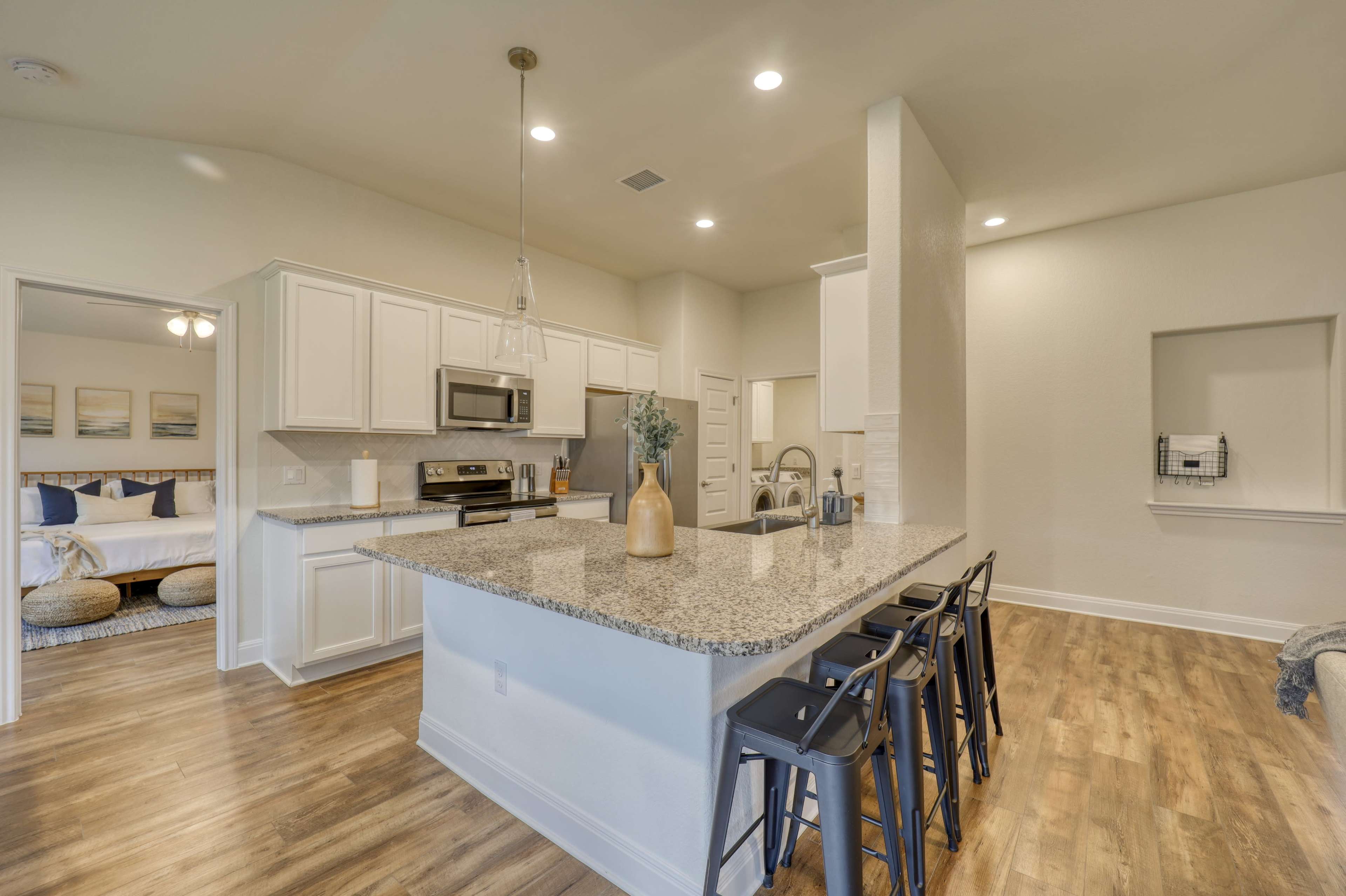 The image shows a modern kitchen with white cabinetry, a large granite island, and bar stools, leading into a connected living area.
