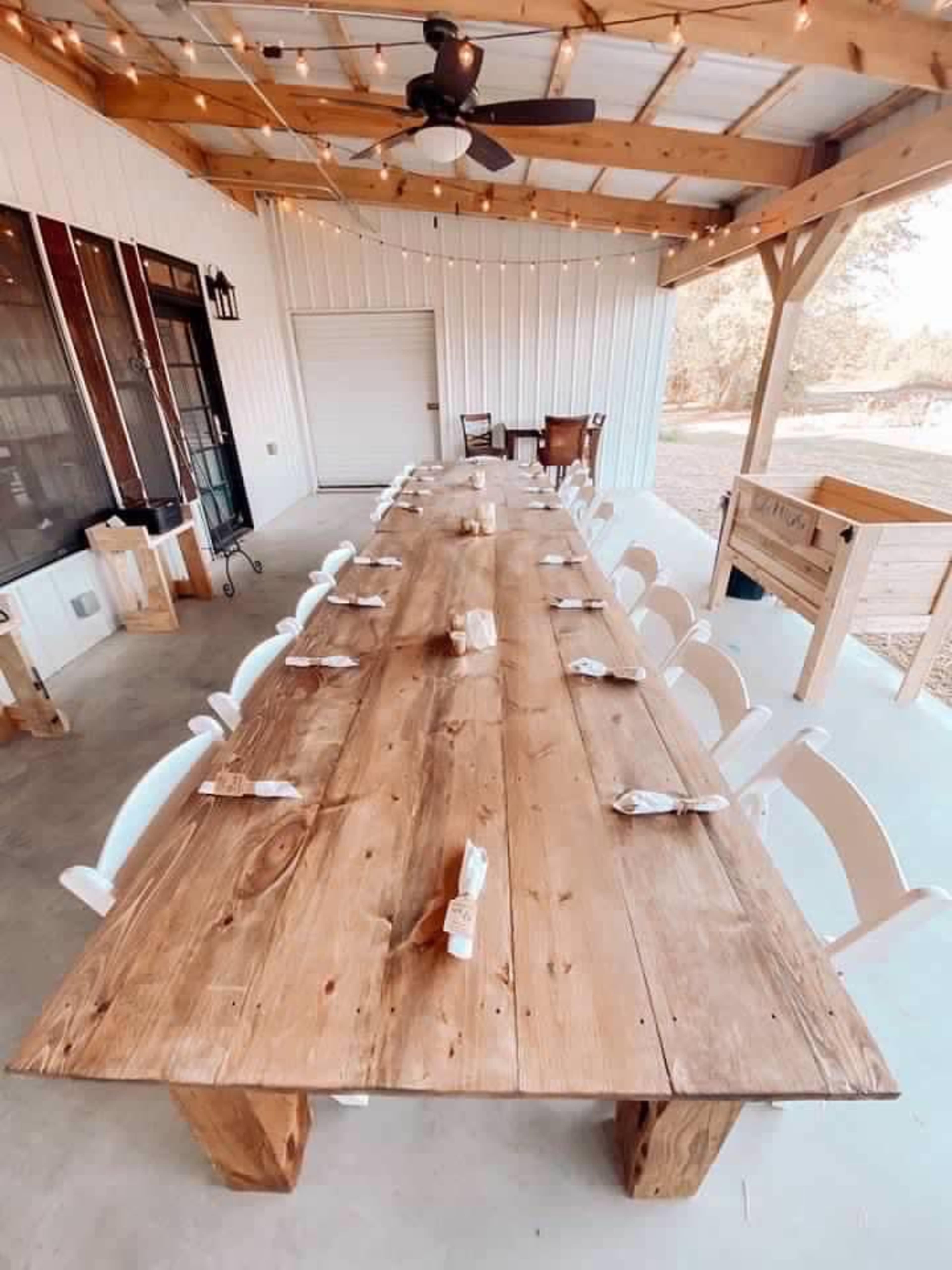 A long wooden table is set for a meal, surrounded by white chairs, beneath a wooden shelter with a ceiling fan.