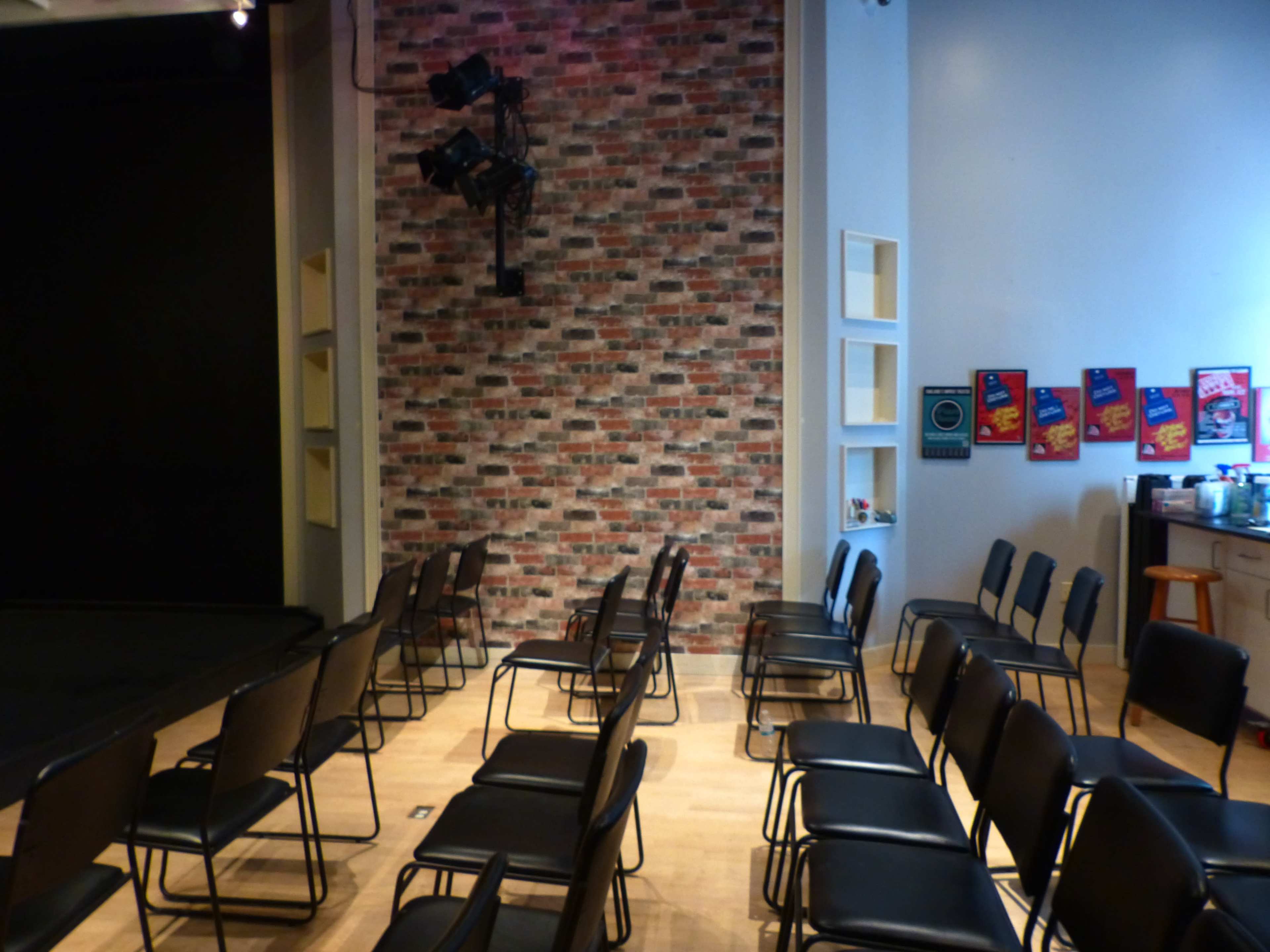 A room with a brick-patterned wall, rows of black chairs arranged facing a small stage.