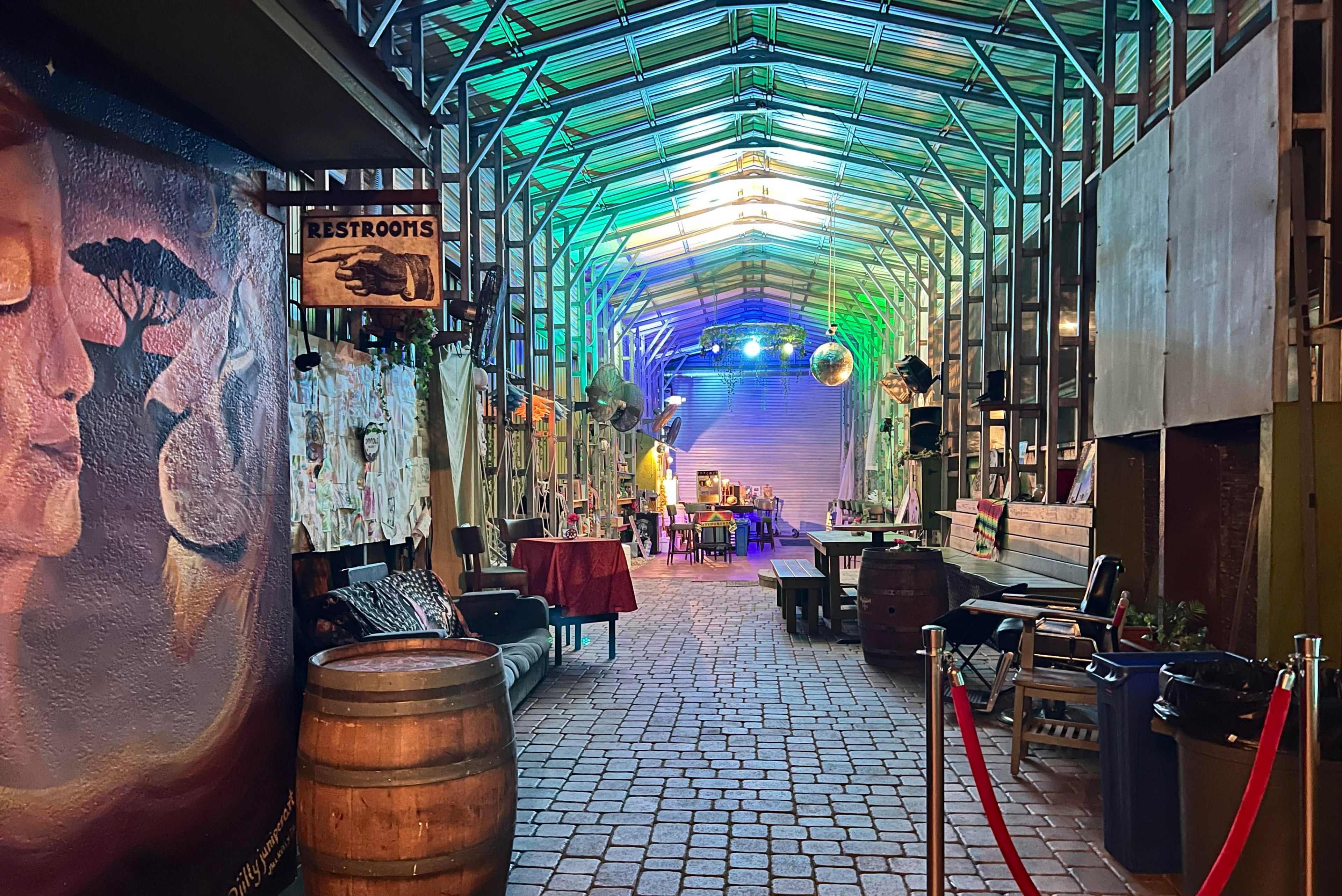A brightly lit corridor lined with benches and decorative elements, featuring a barrell at one end and a sign for restrooms.