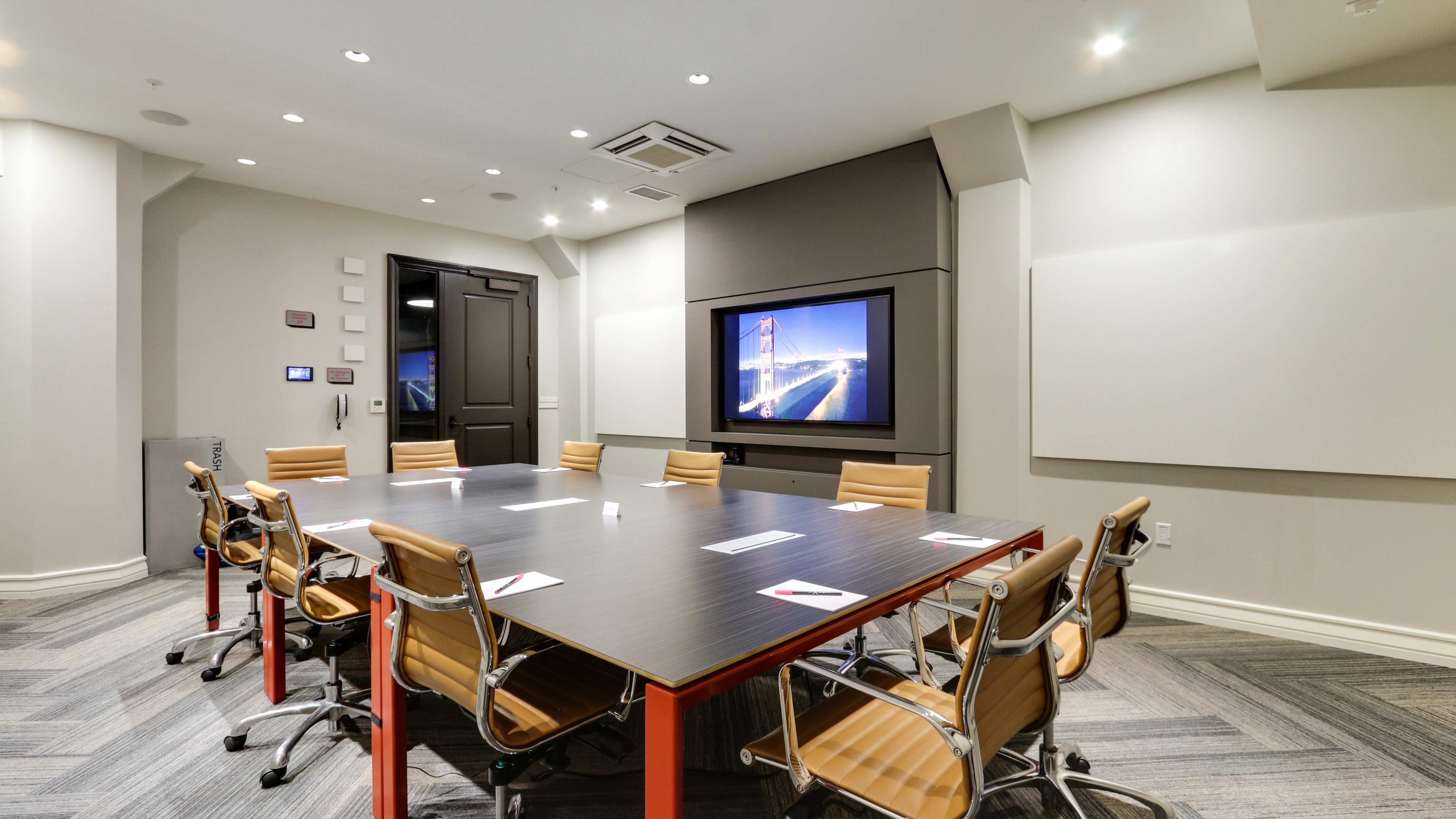 A modern conference room features a large rectangular table surrounded by ergonomic chairs, with a wall-mounted screen displaying an image.