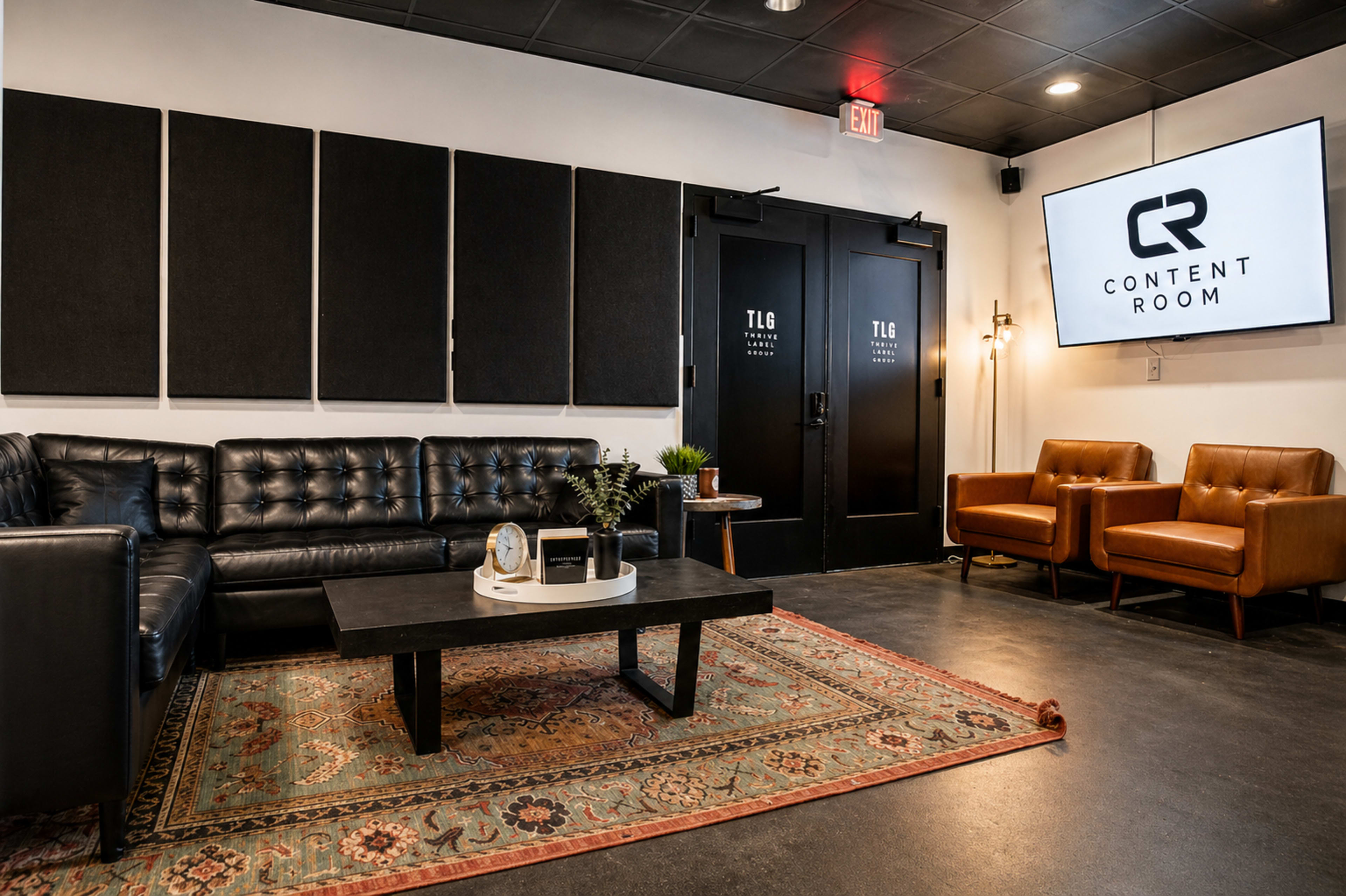 The image shows a modern cozy lounge area featuring a black leather sofa, two brown armchairs, a coffee table on a decorative rug, and a television displaying the words "CONTENT ROOM" on the wall.