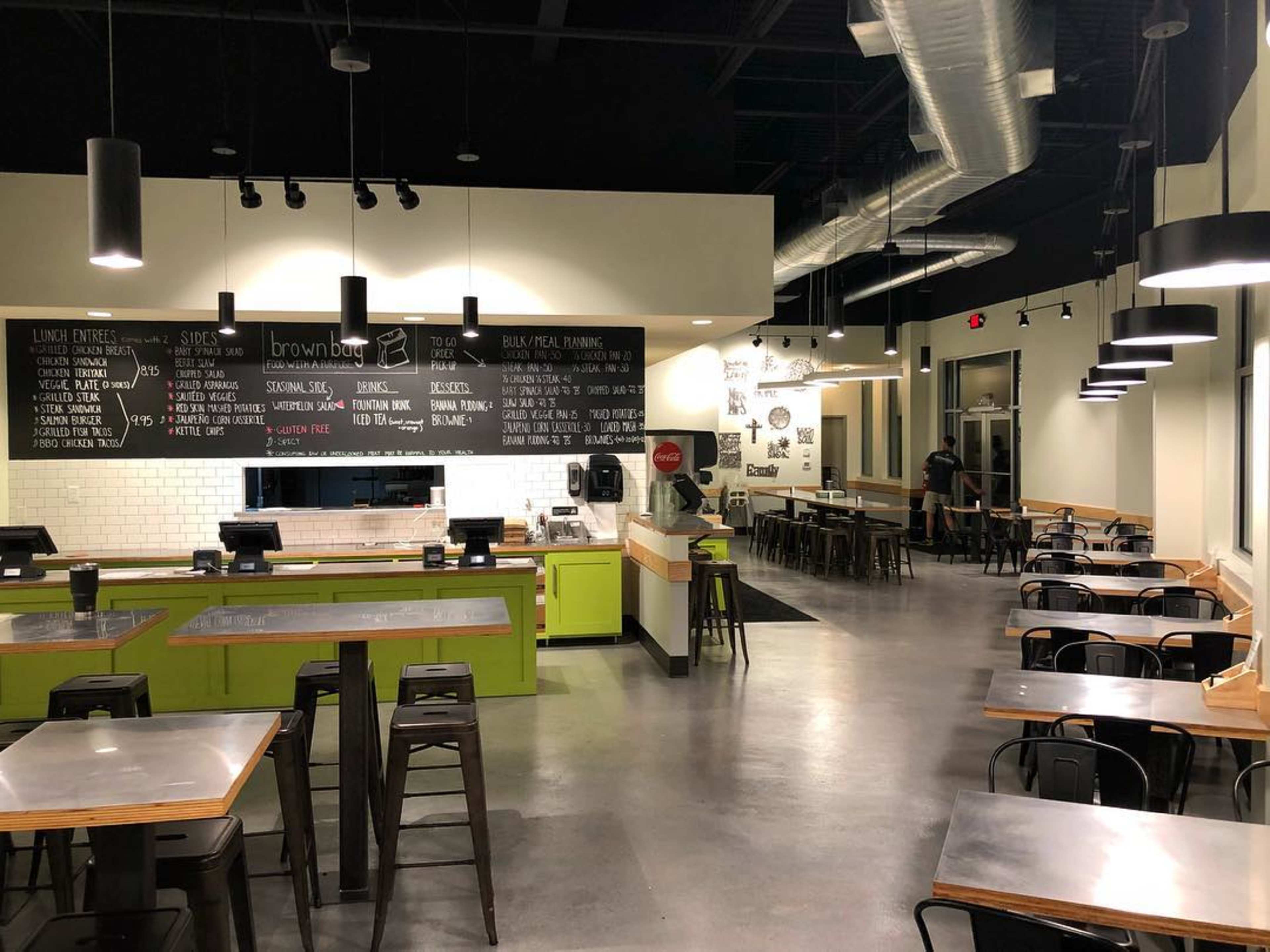 A modern restaurant interior with green accents, a menu written on a black wall, and rows of metal and wooden tables.