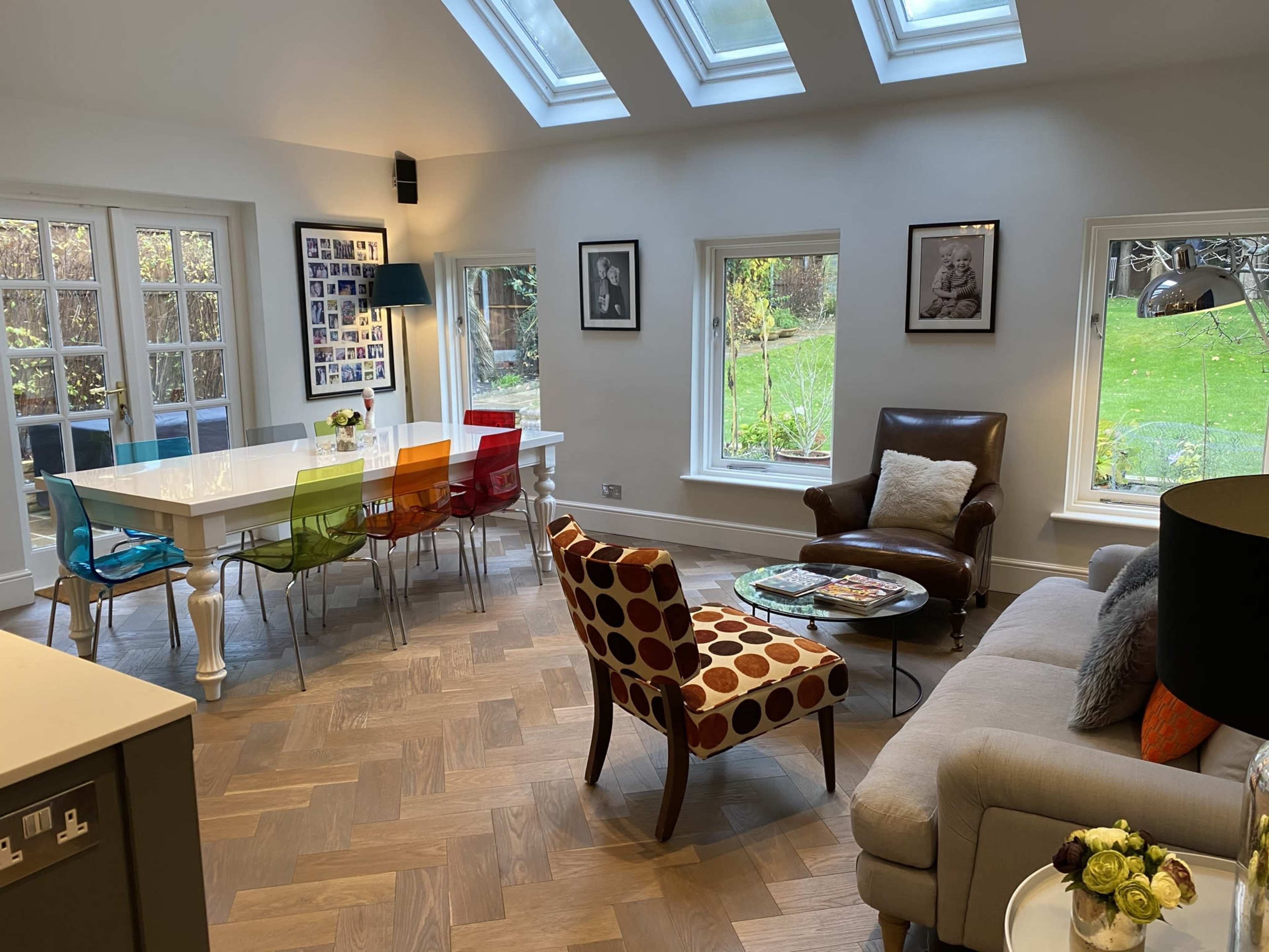 A modern living space with a dining table surrounded by colorful chairs, a comfortable armchair and sofa, and large windows letting in natural light.