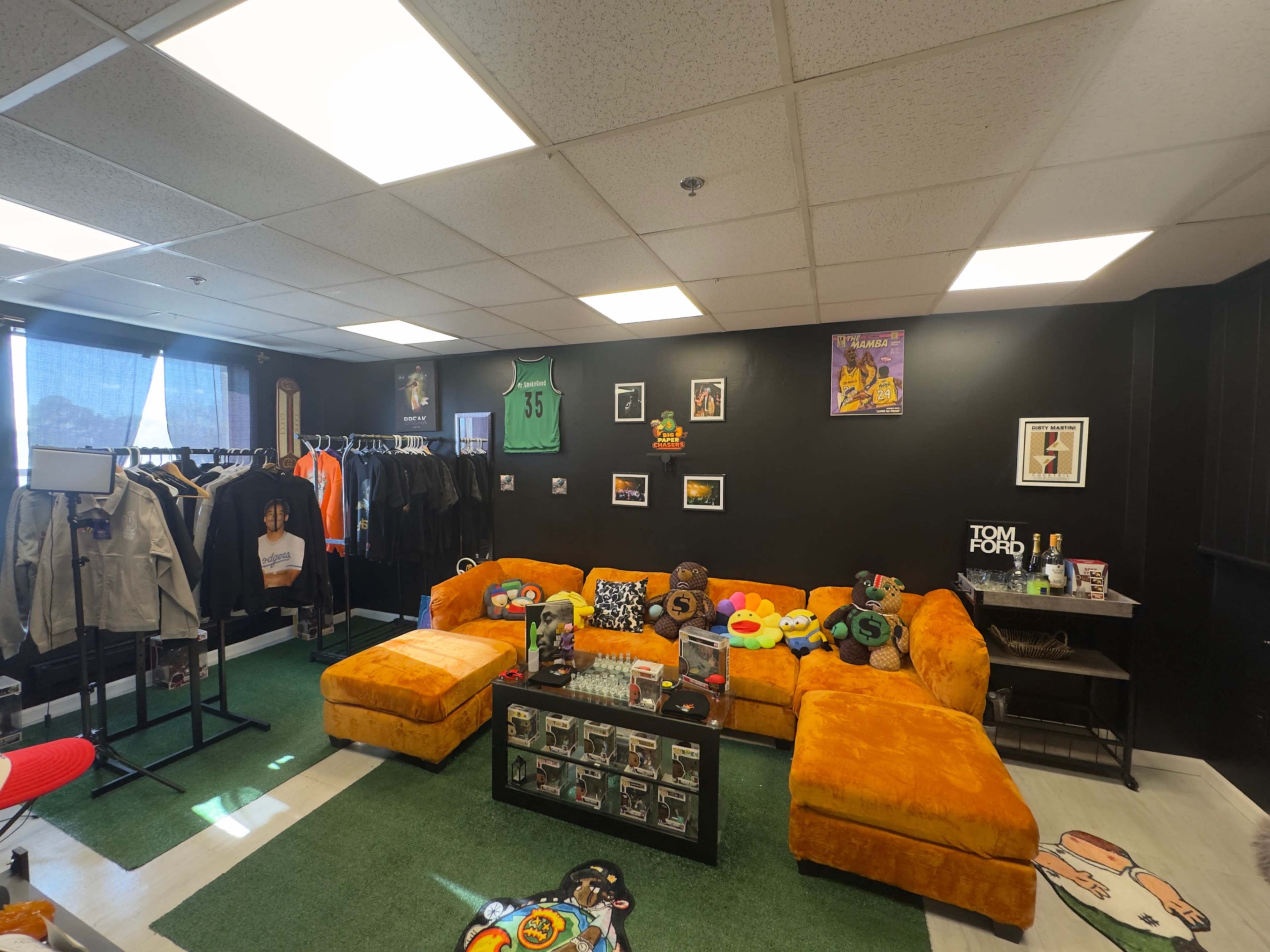 The image shows a vibrant room featuring a large orange sectional couch, clothing racks with various garments, and a black wall decorated with framed pictures and art.
