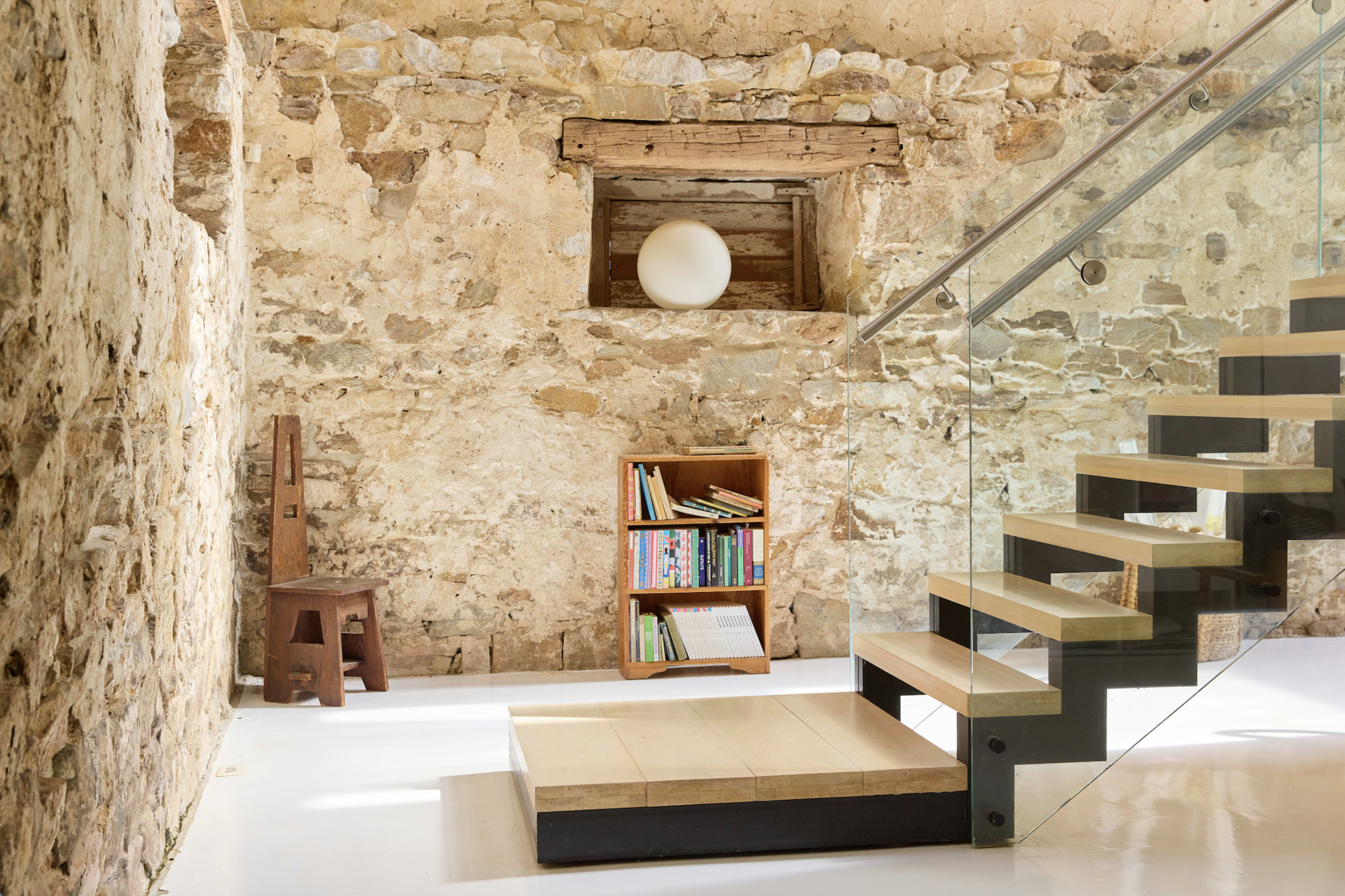 The image shows a modern interior space with a stone wall, a wooden shelf filled with books, a minimalist white orb in a window, and a staircase with glass railing.