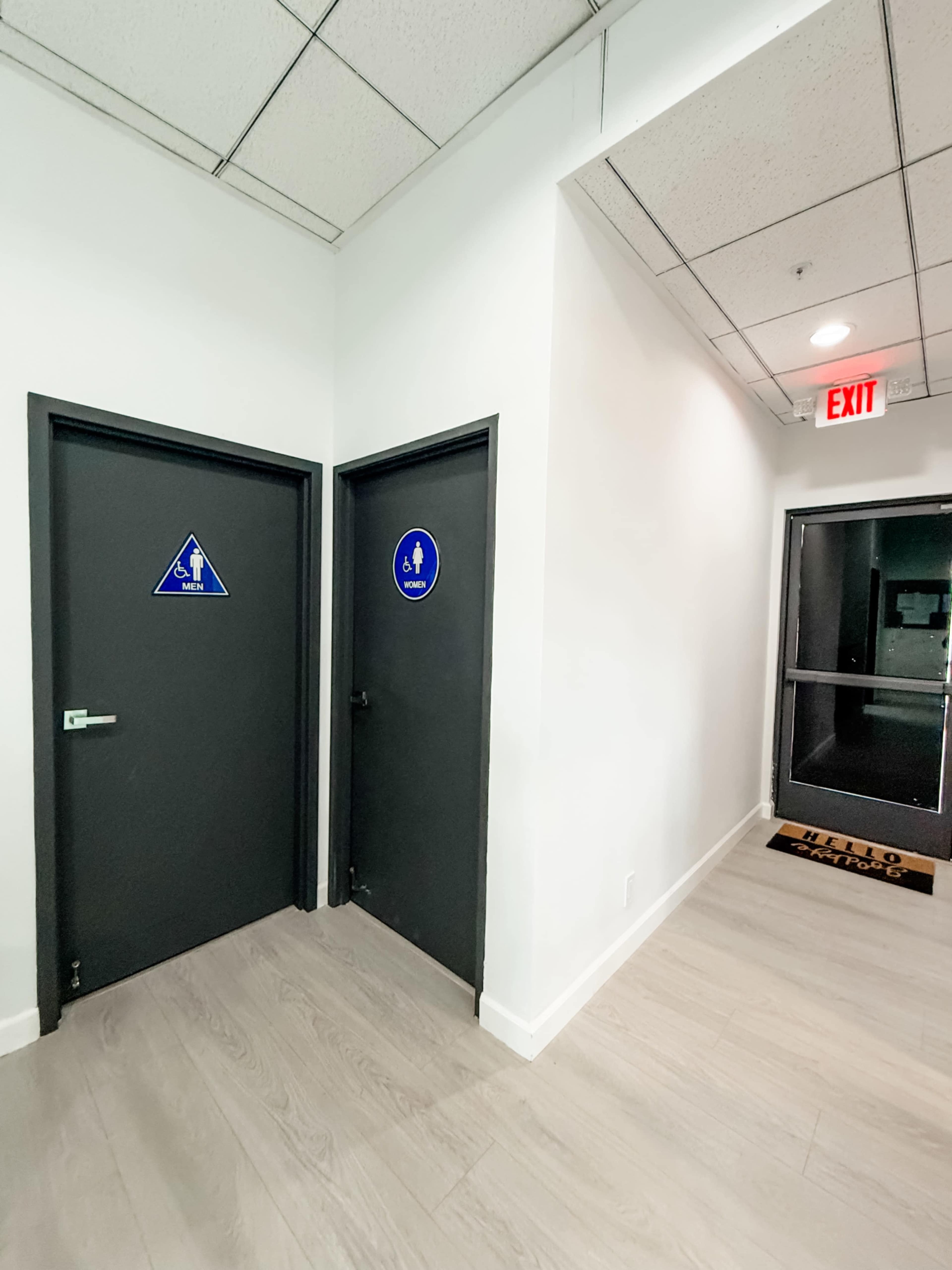The image shows a hallway with two restroom doors on the left, one labeled for women and the other for men, along with an exit door on the right.