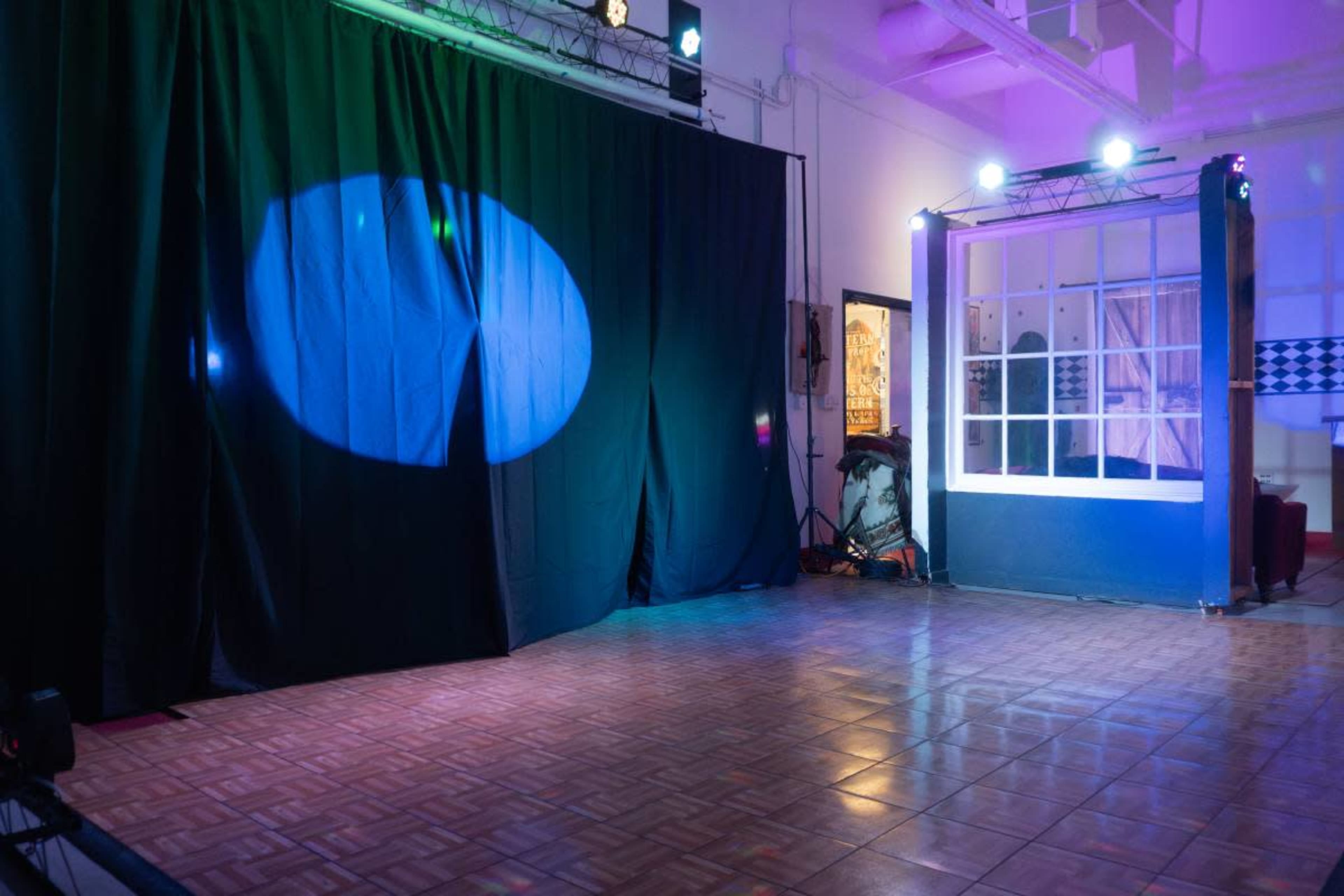 The image shows a performance space with a black curtain on one side and colorful stage lighting illuminating a wooden floor.