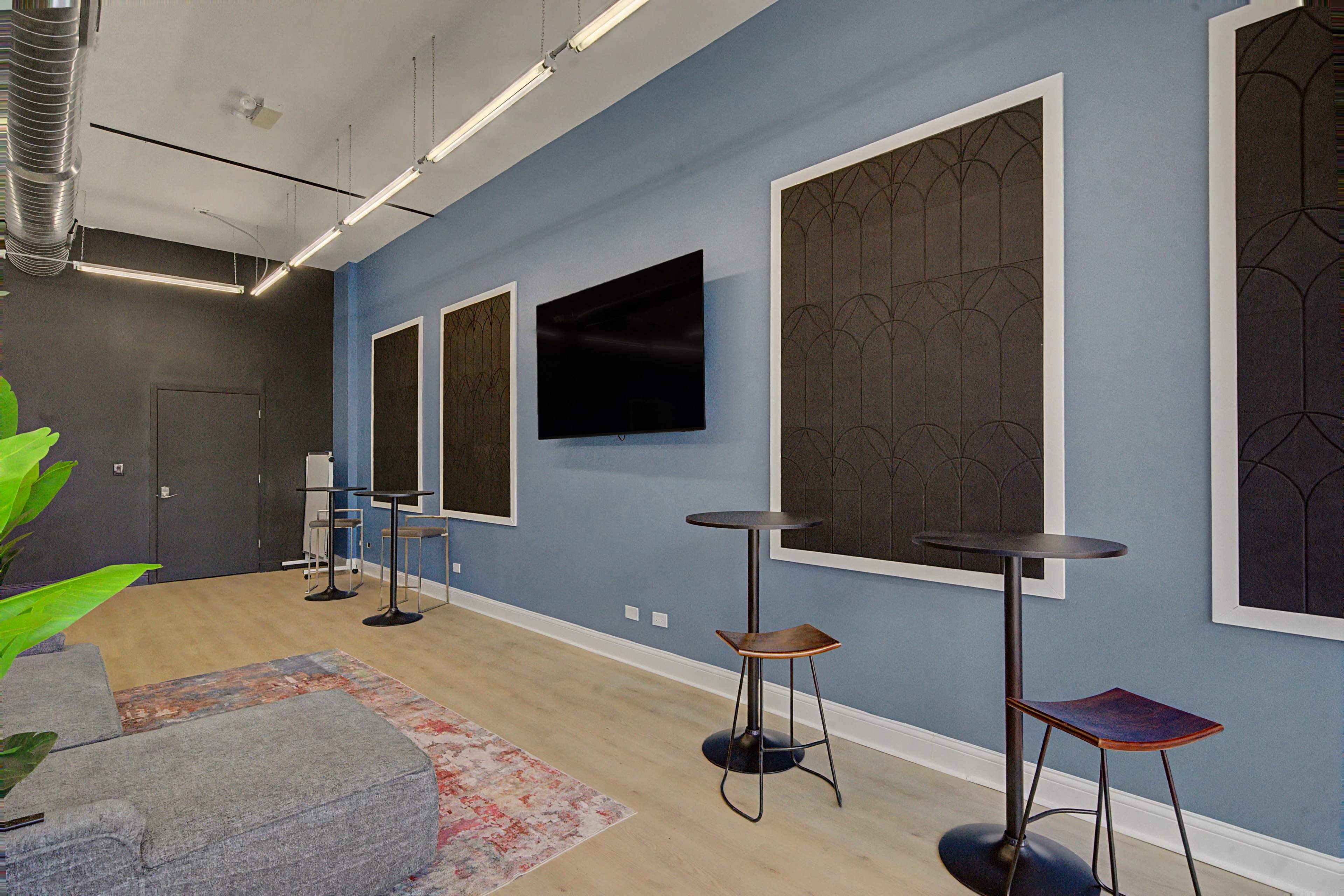 The image shows a modern office space with a gray sofa, two black tables, and three large framed wall panels, alongside a wall-mounted television and greenery.