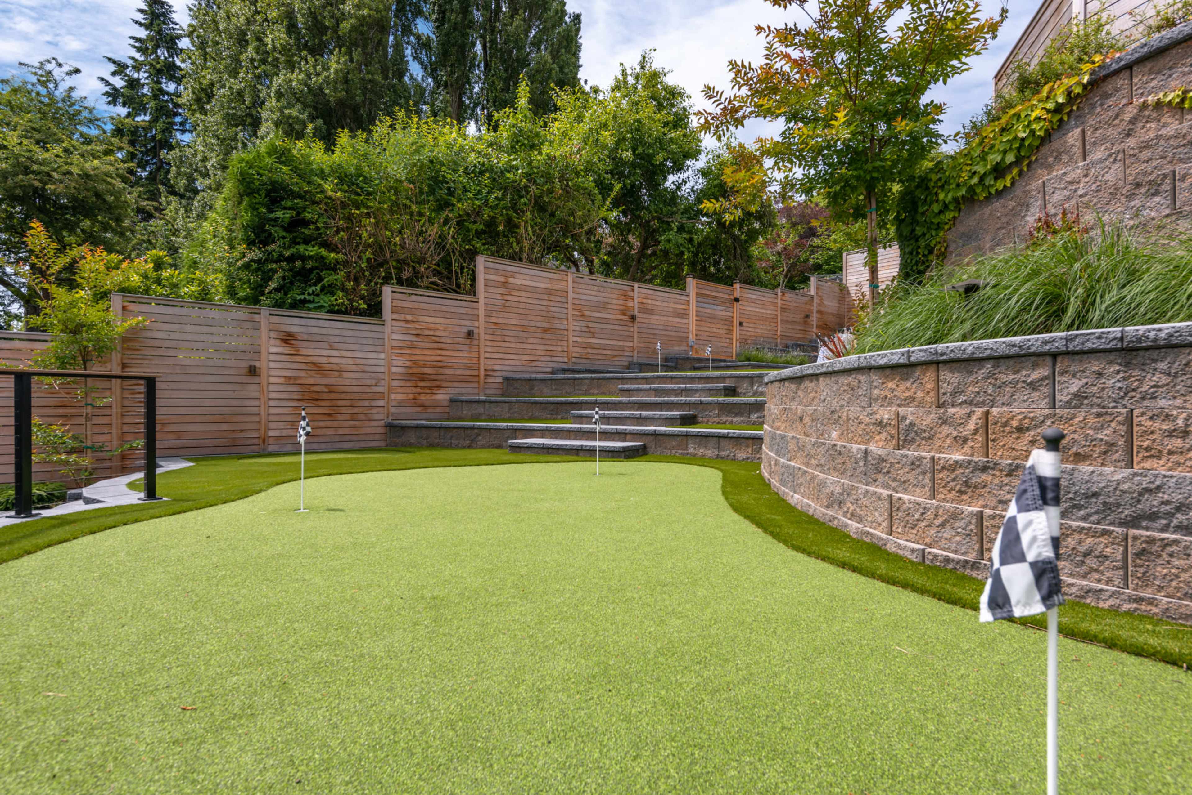 A landscaped putting green with a series of steps and wooden fencing, surrounded by greenery.