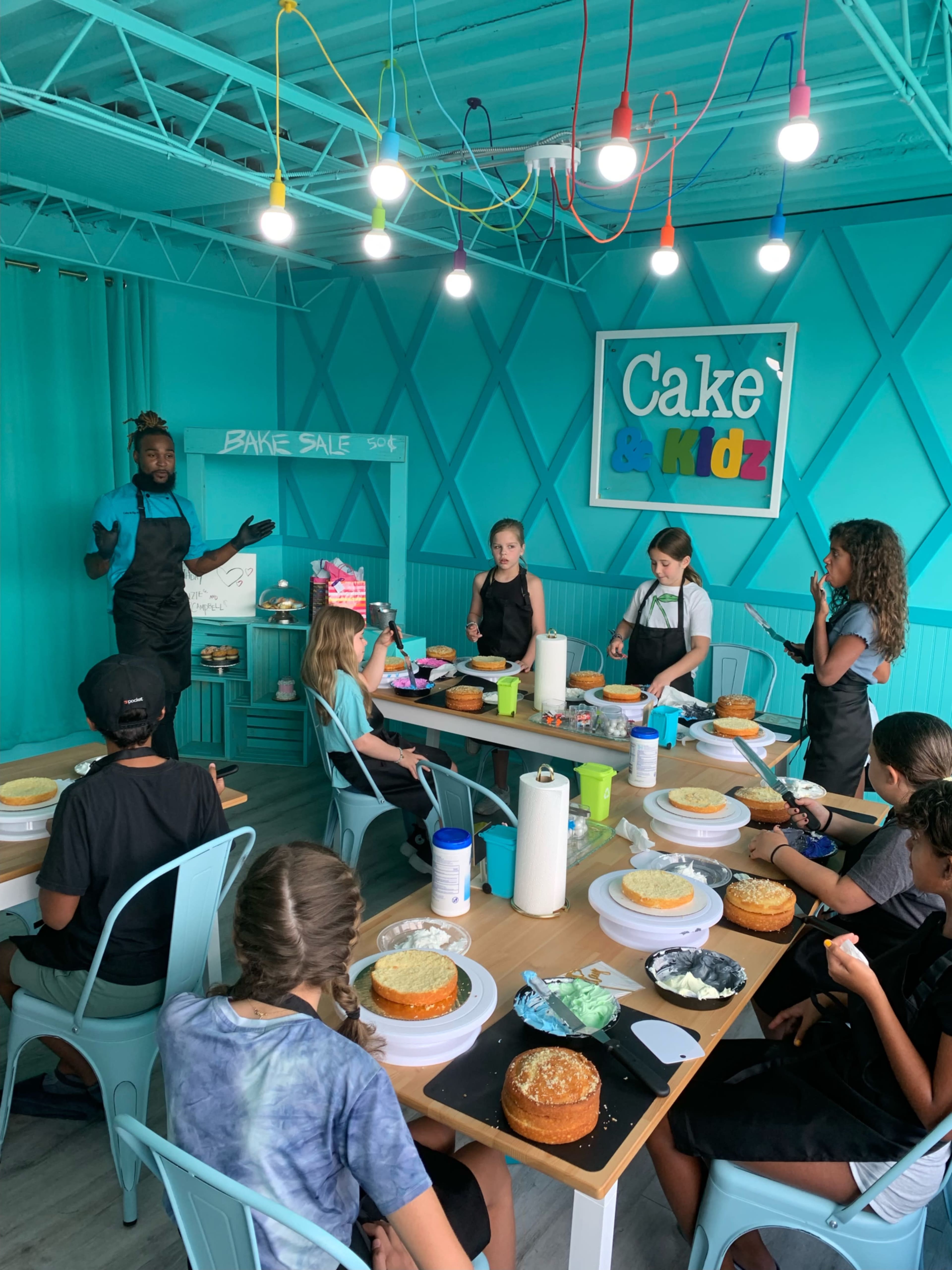 A group of children participate in a baking class, each working on decorating layered cakes in a brightly colored room.