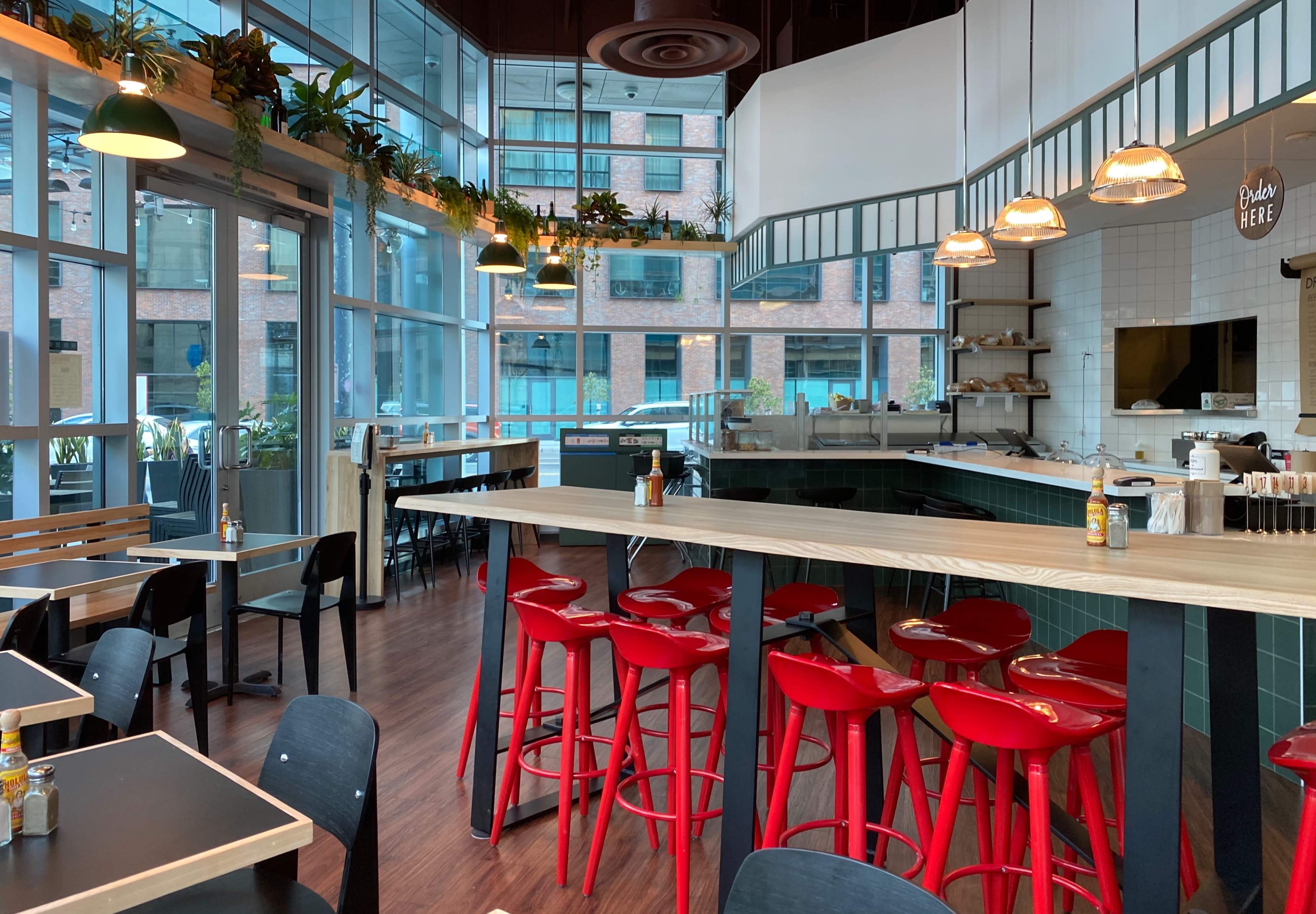 A modern café interior with large windows, wooden tables, red bar stools, and greenery along the walls.