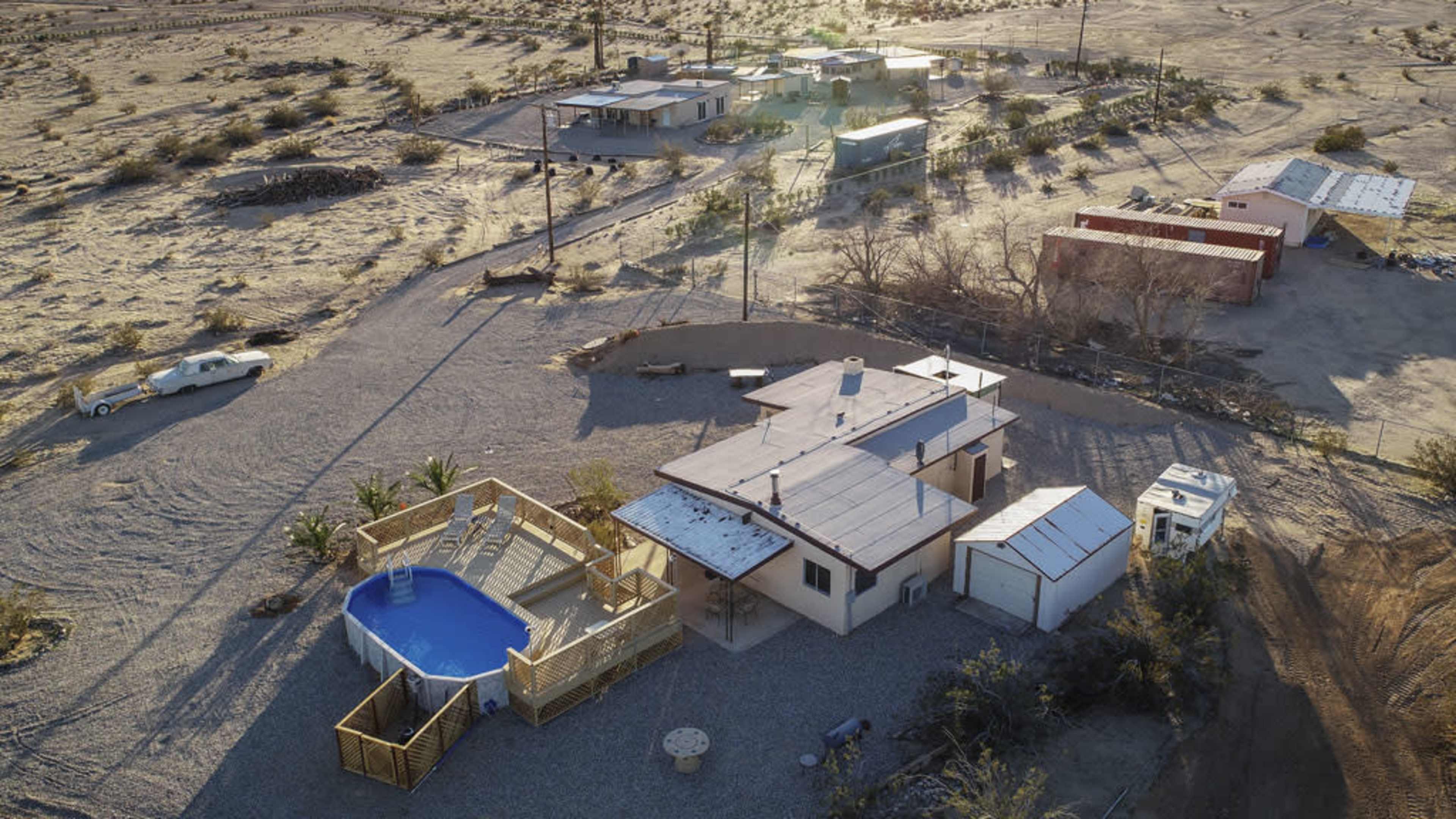 A cluster of buildings in a desert landscape, including a house with a pool, surrounded by gravel and sparse vegetation.