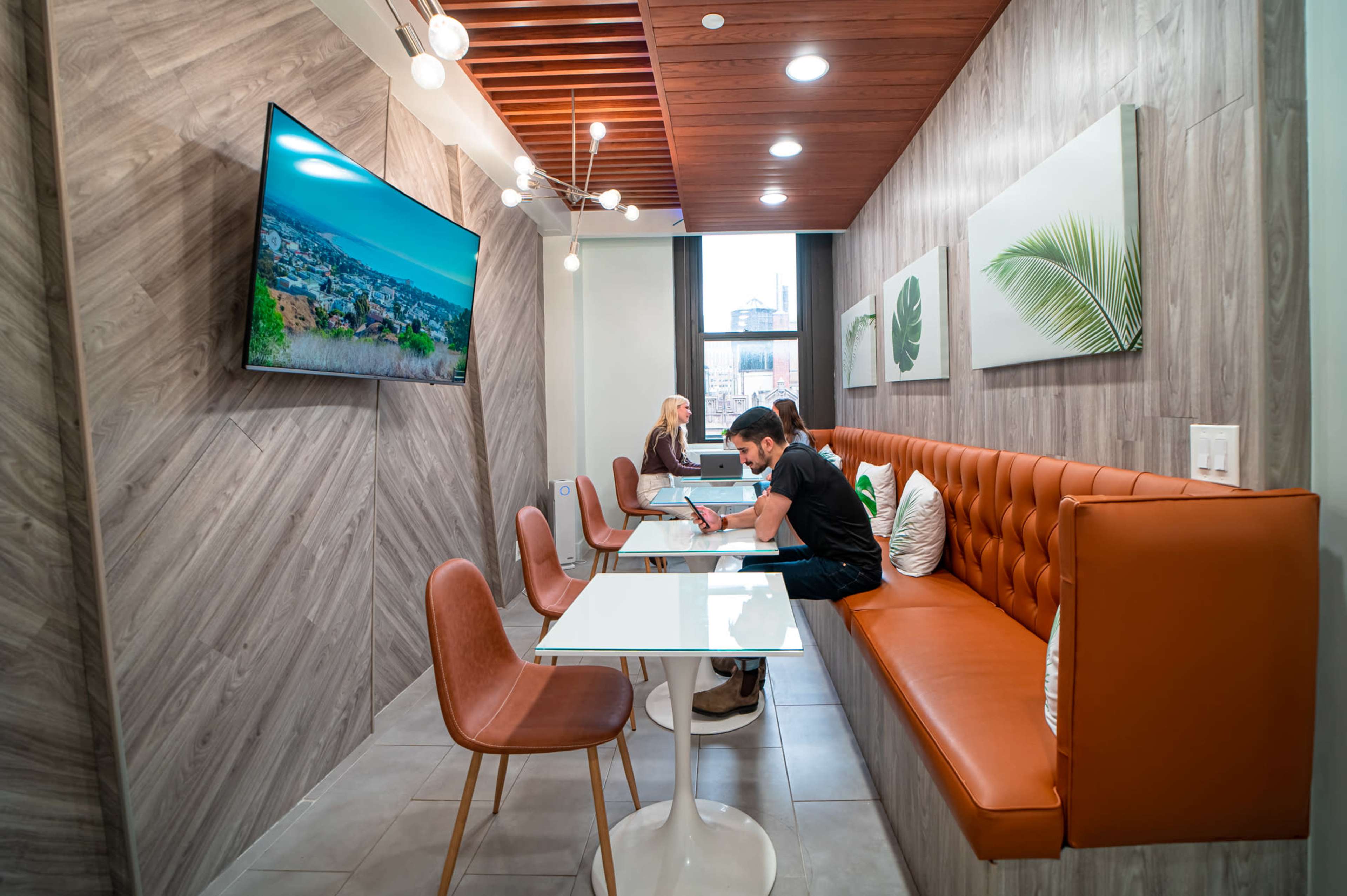 A modern, minimalistic room with a seating area featuring a leather bench and tables, where two people are engaged in work while a television displays a landscape.
