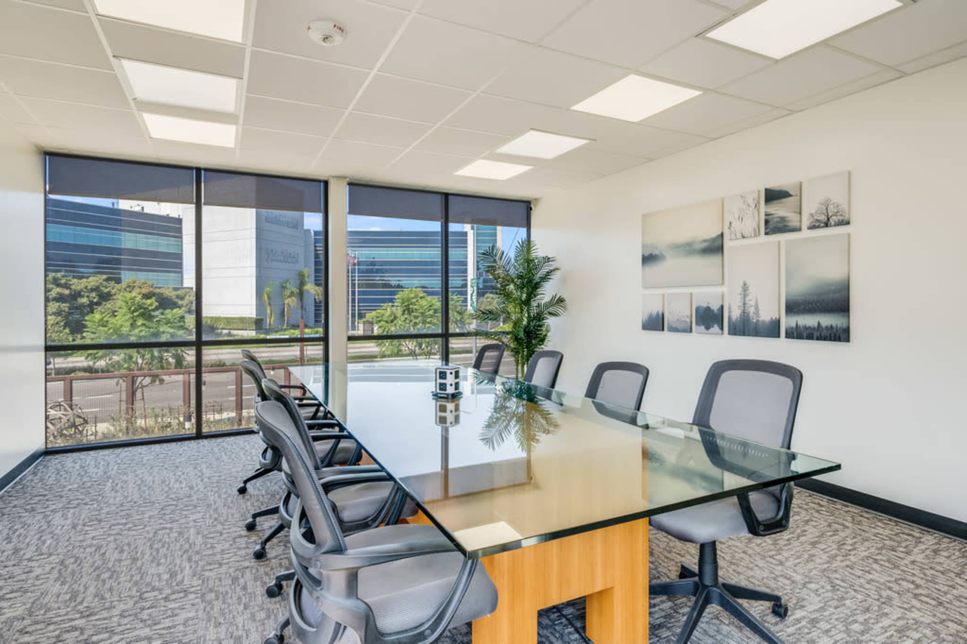 A modern conference room with a large glass table surrounded by several office chairs, and large windows with a view of a landscaped area outside.