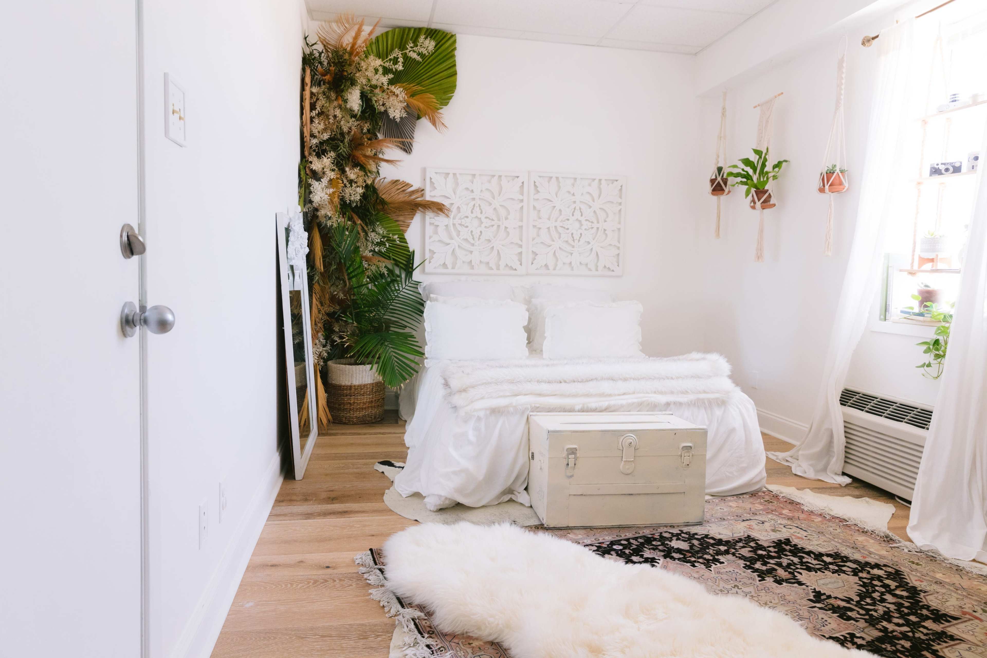 A bright bedroom features a white bed with plush bedding, a decorative wall panel, and plants arranged near a window, with a vintage chest and a large mirror in the corner.