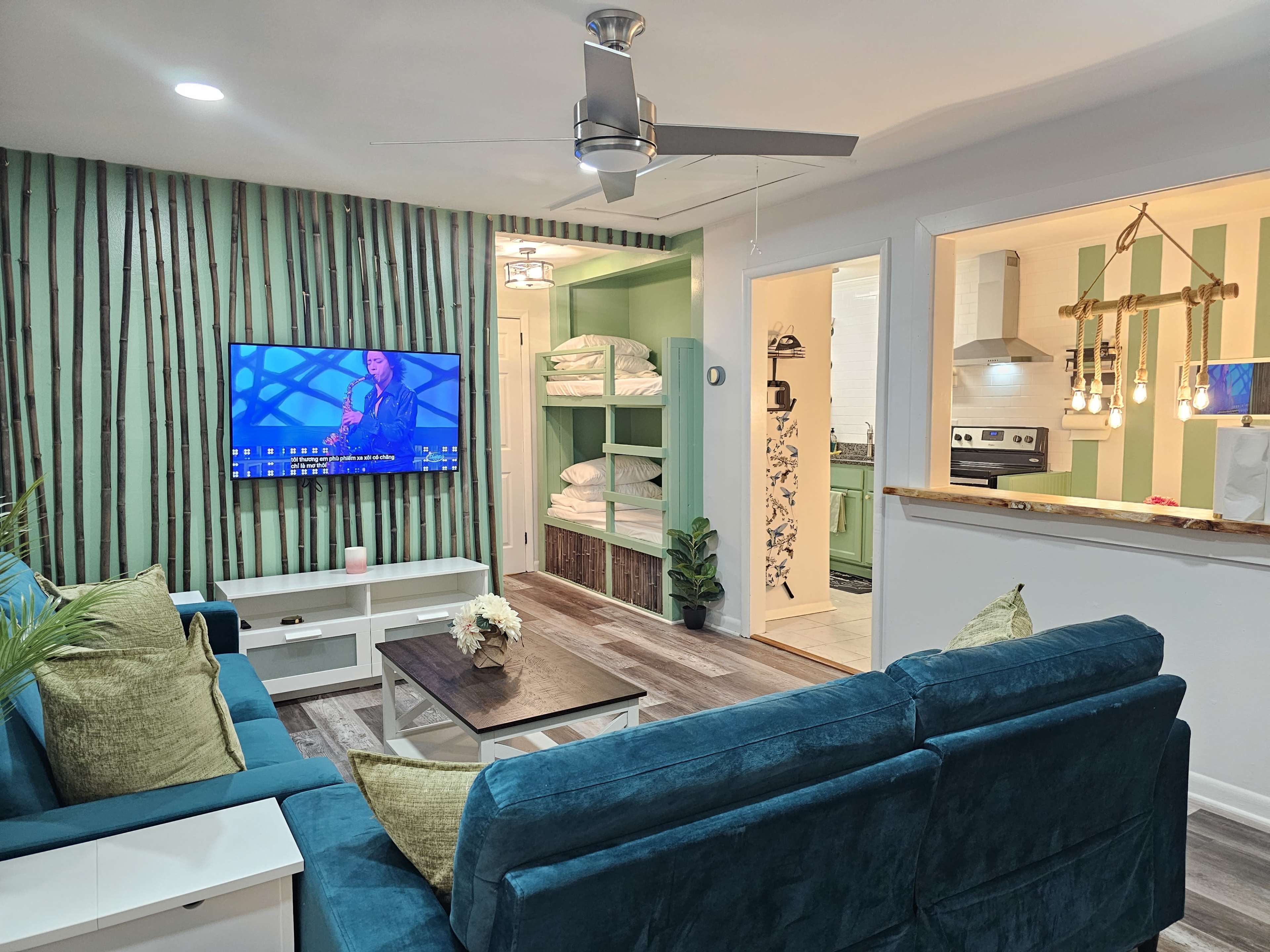 The image shows a modern living room with a teal sectional sofa, a wall-mounted TV, and a visible open kitchen area with stacked white towels and a decorative light fixture.