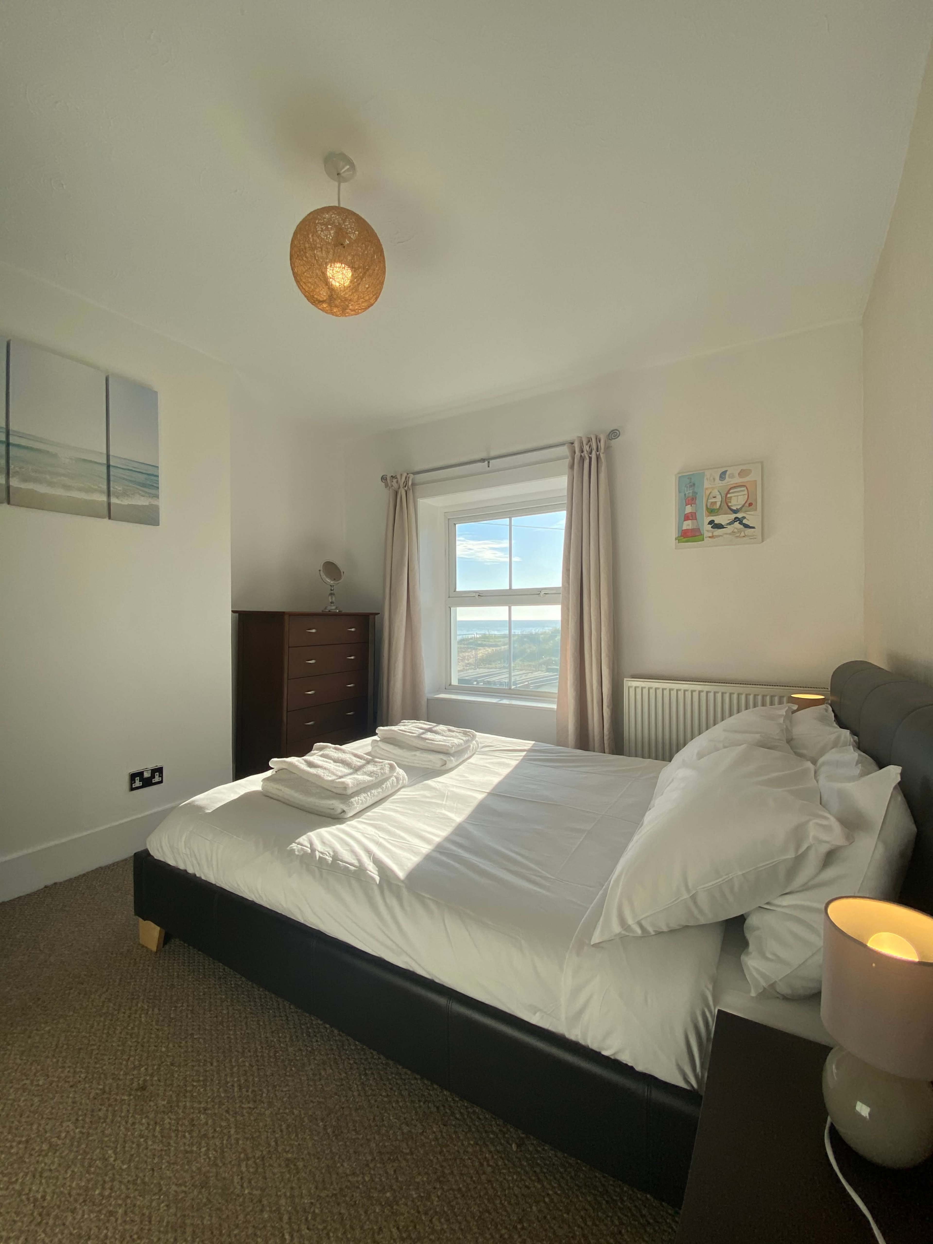 A cozy bedroom features a large bed with white linens, a wooden dresser, and a window offering a view of the outside landscape.
