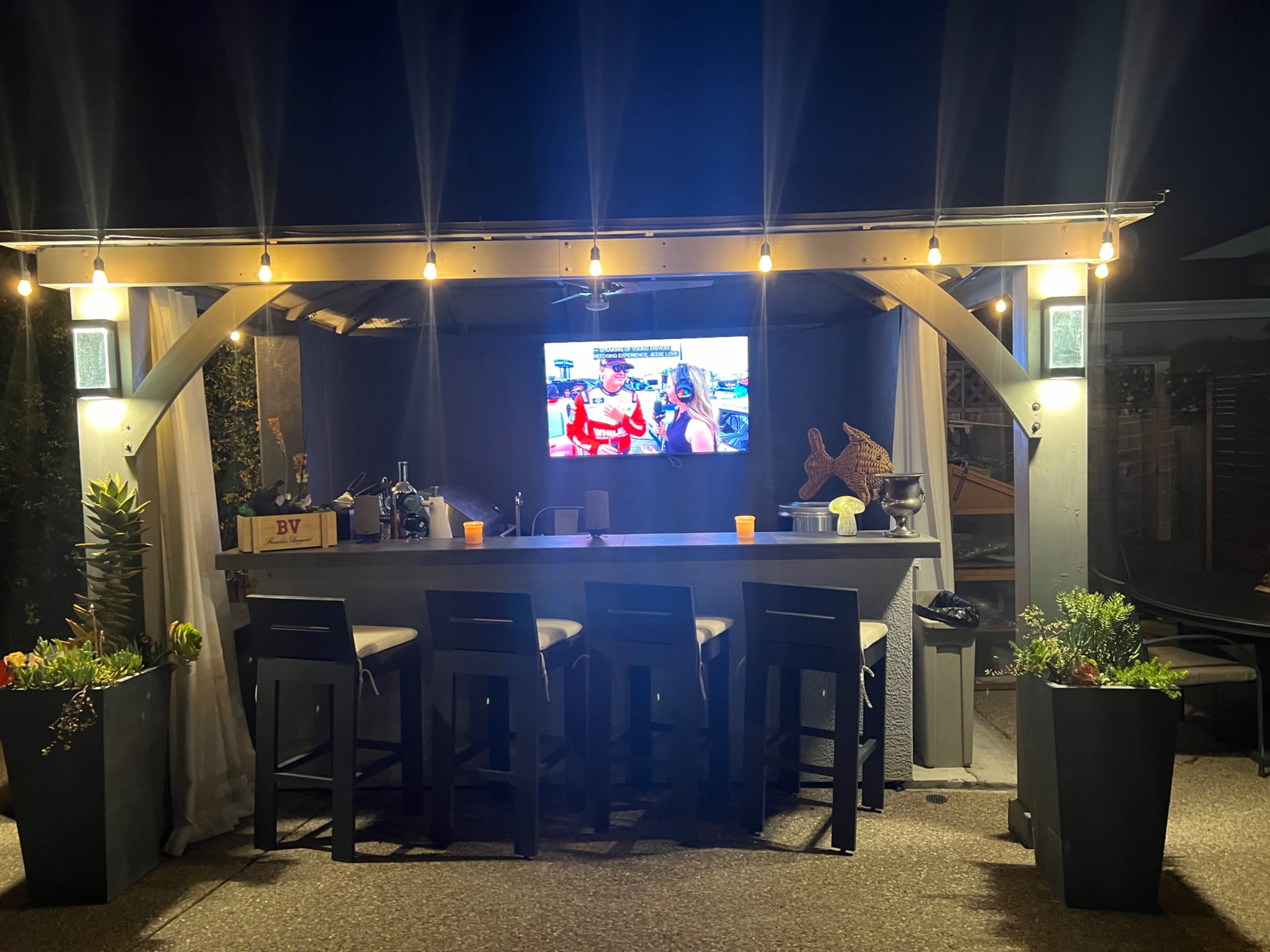 A covered outdoor bar with high stools, illuminated by string lights, features a television mounted on the wall displaying a show, surrounded by potted plants.