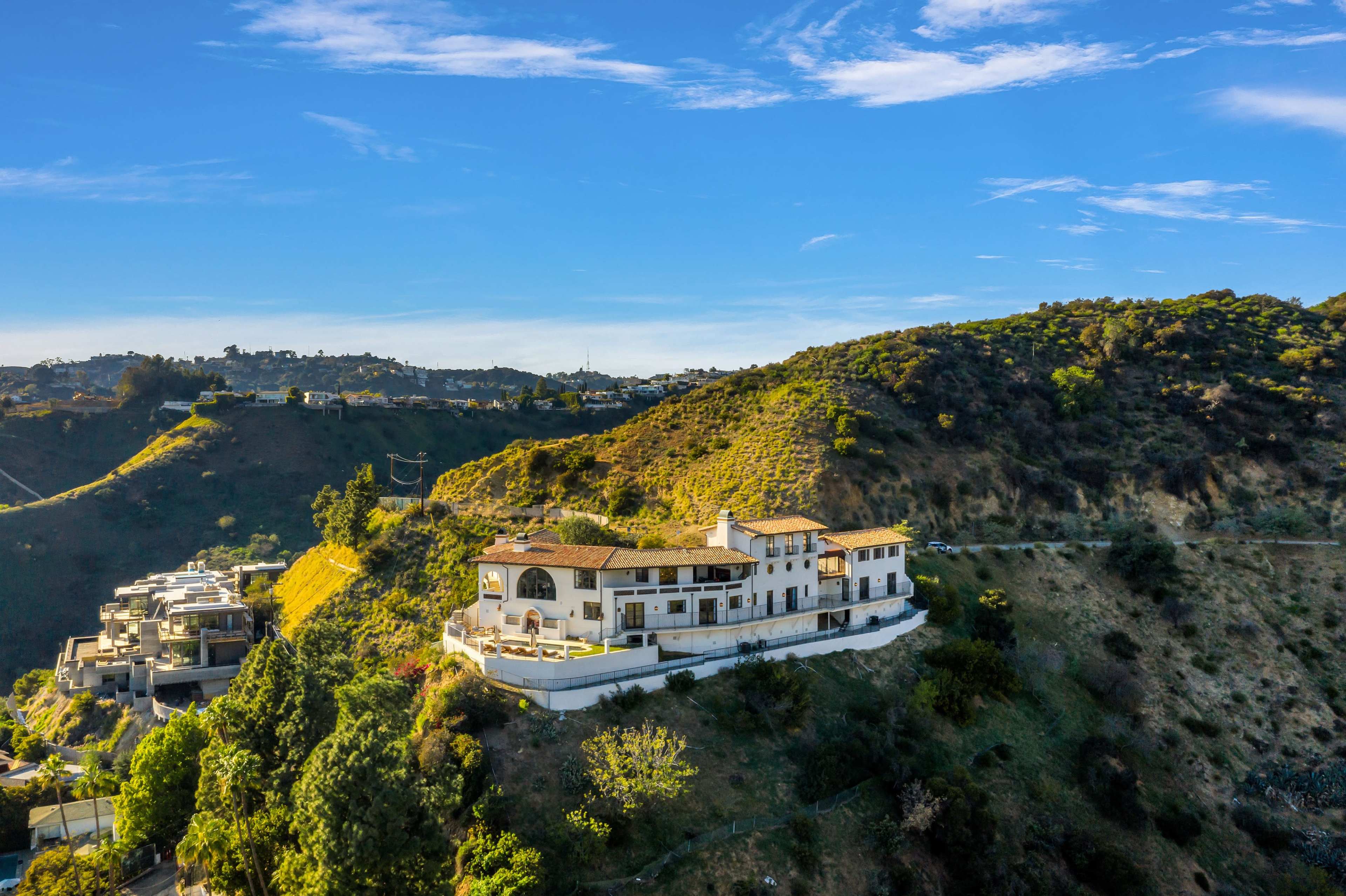A large, multi-level house stands on a hillside, surrounded by greenery and overlooking a valley.