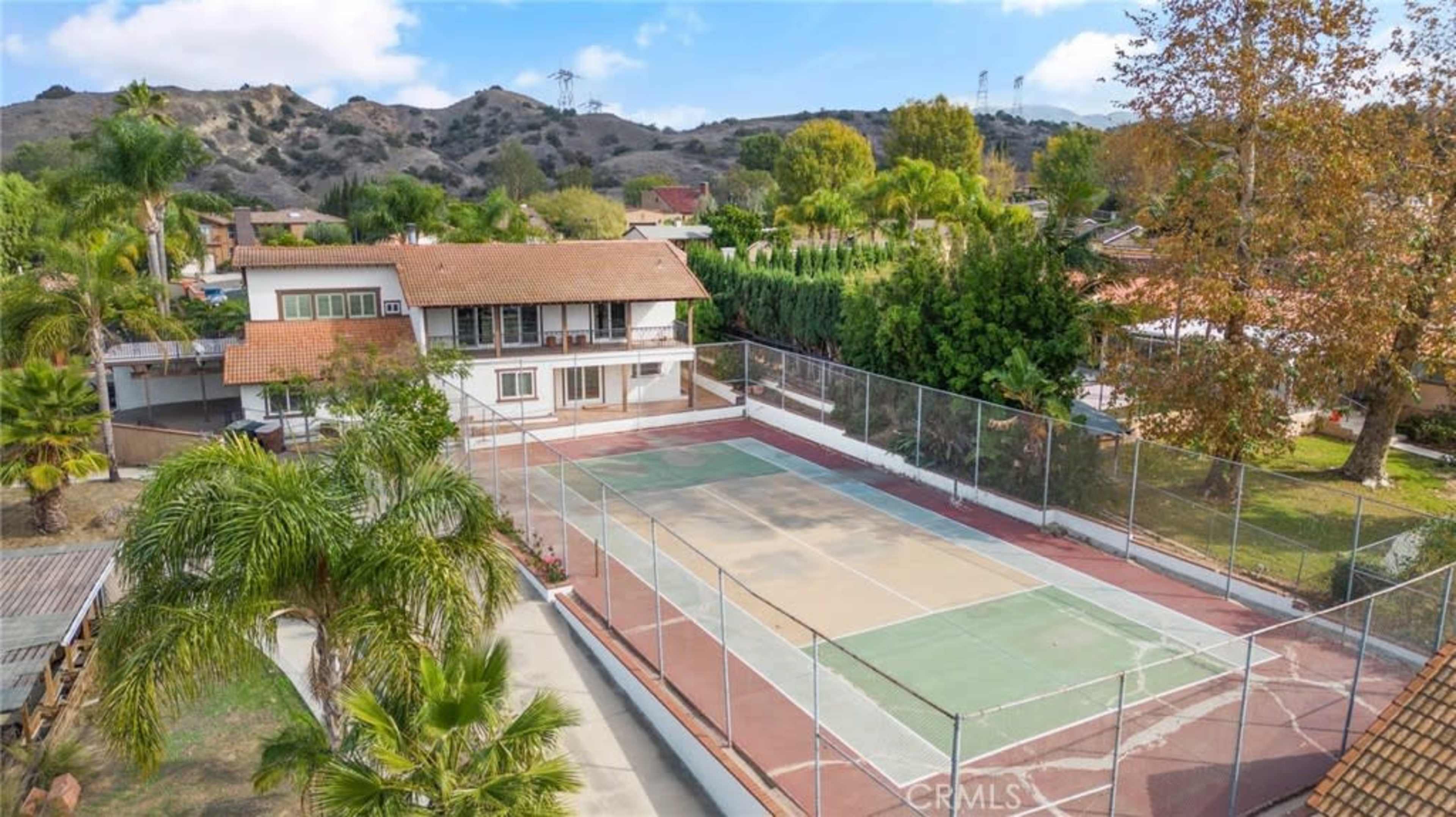 The image shows a residential property with a fenced tennis court surrounded by trees and hills in the background.