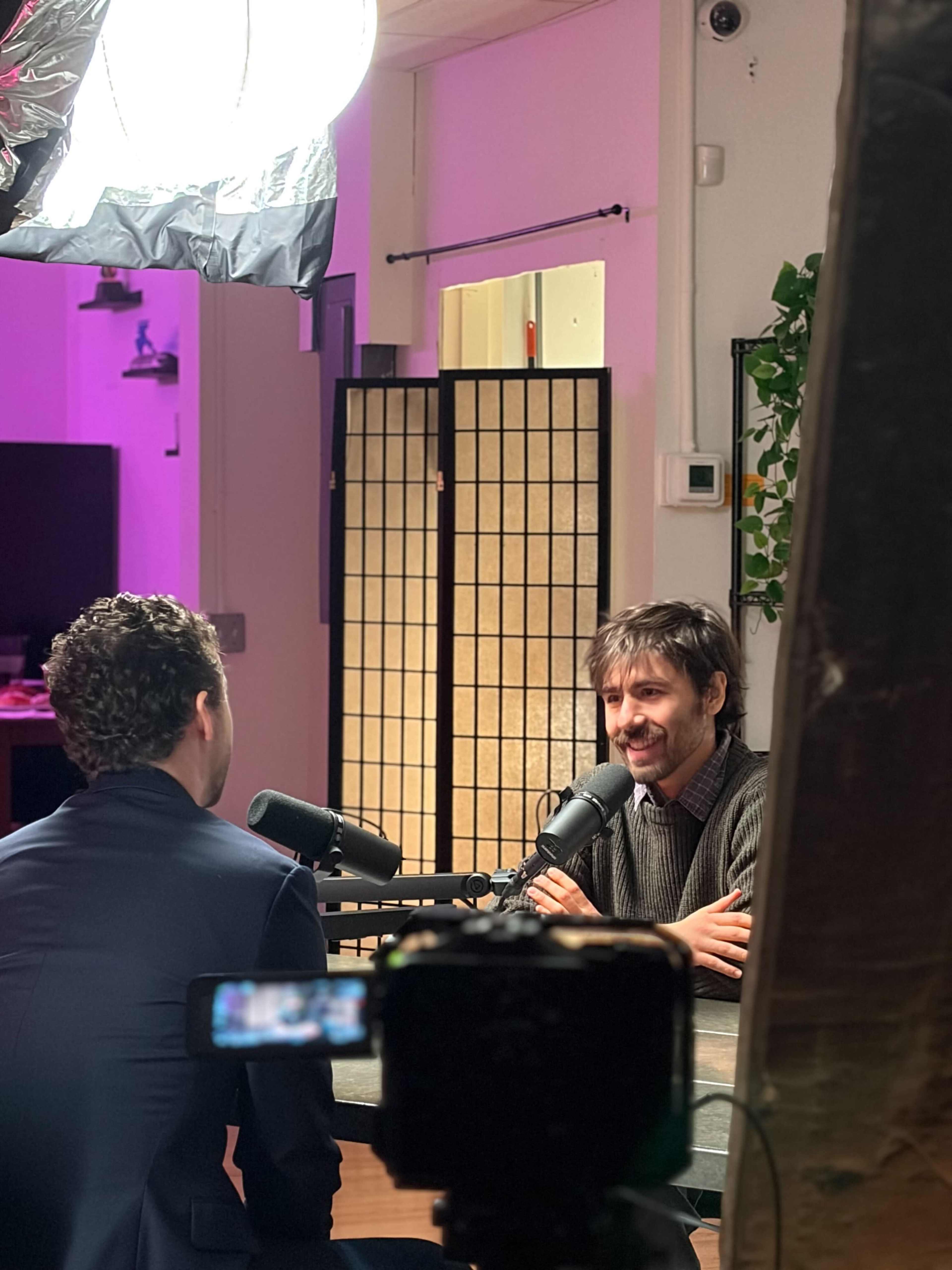 Two men are seated at a table with microphones, engaged in a discussion in a well-lit studio with a soft pink backdrop.