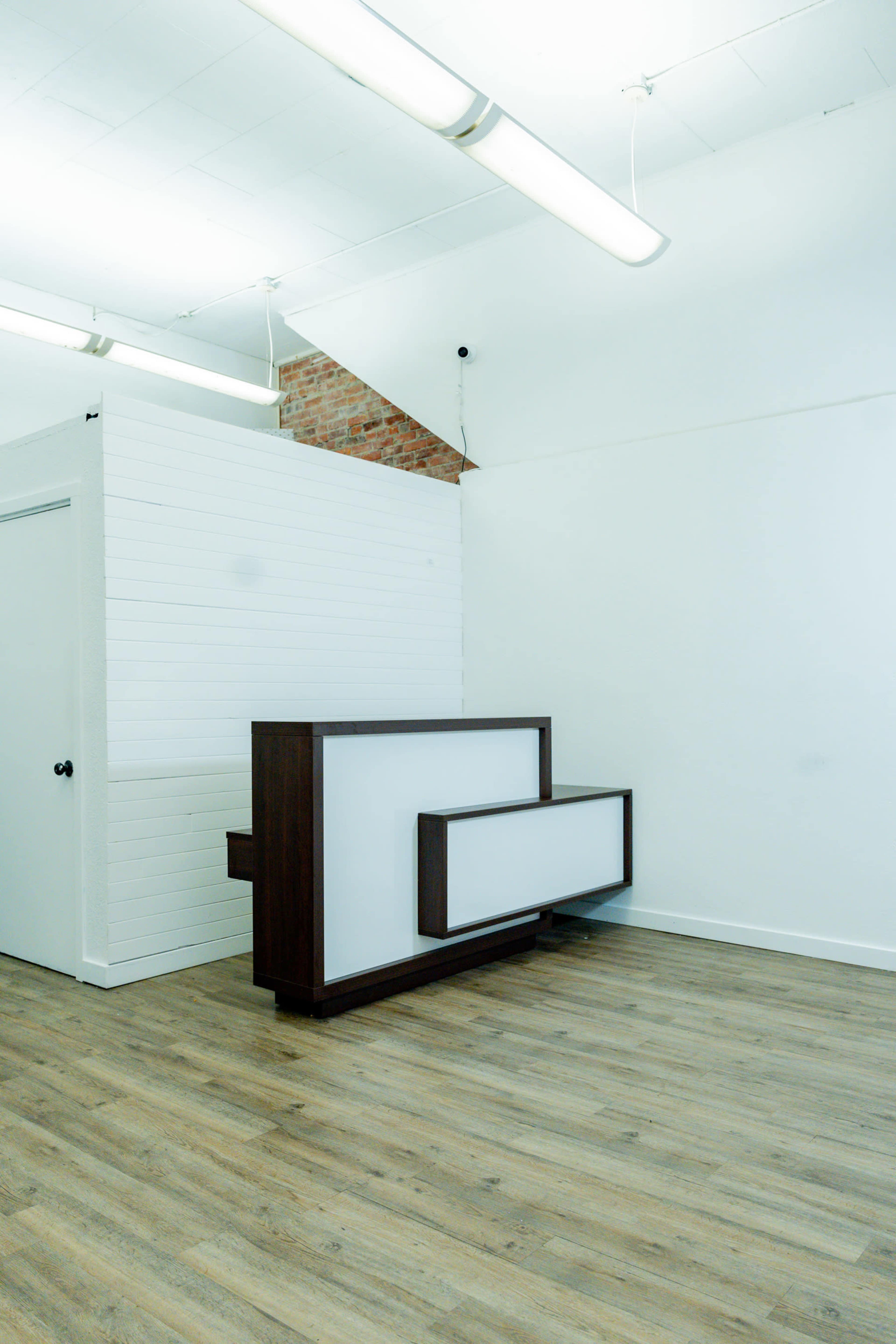 A minimalist interior space featuring a wooden reception desk against a white wall with a door and a brick accent.