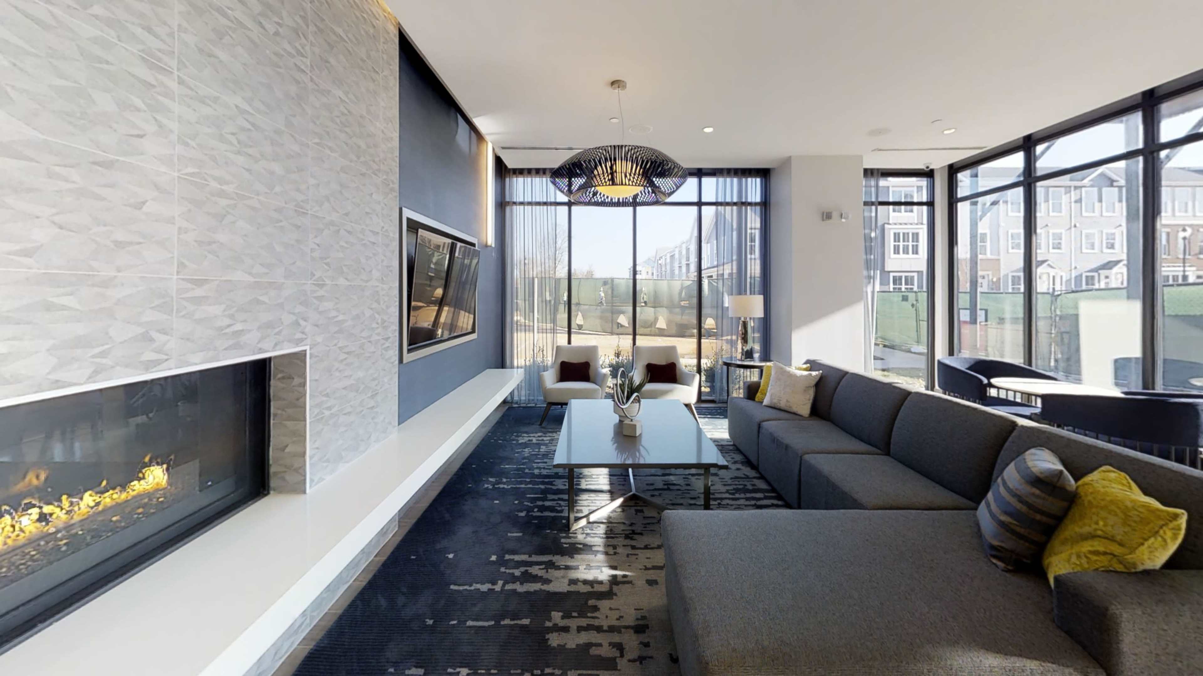 A modern living room with a large sectional sofa, a flat-screen TV mounted on the wall, and a decorative fireplace, all illuminated by natural light through large windows.