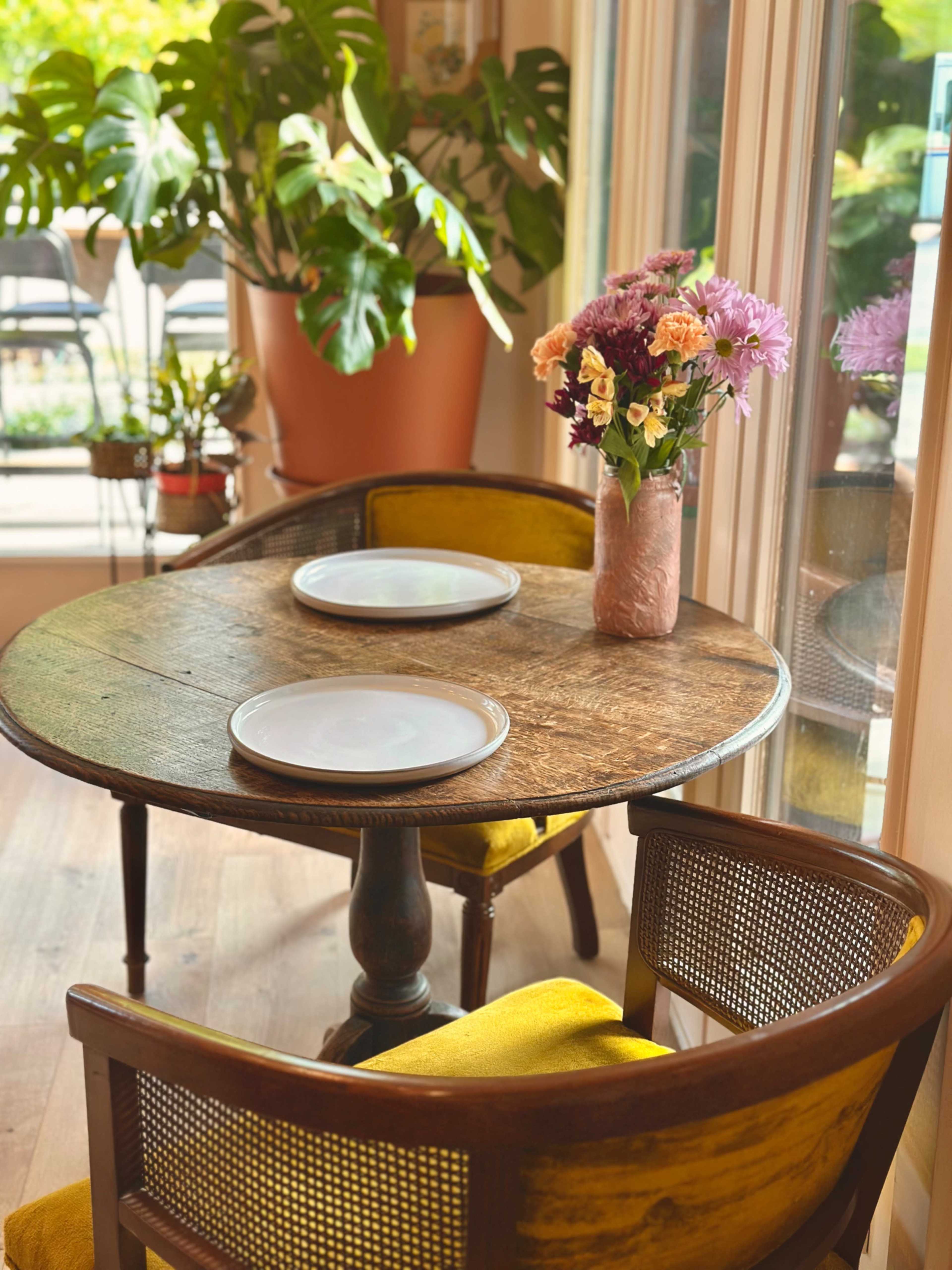 A small round wooden table with two chairs, set with two plates, is placed near a window with a bouquet of flowers in a vase.