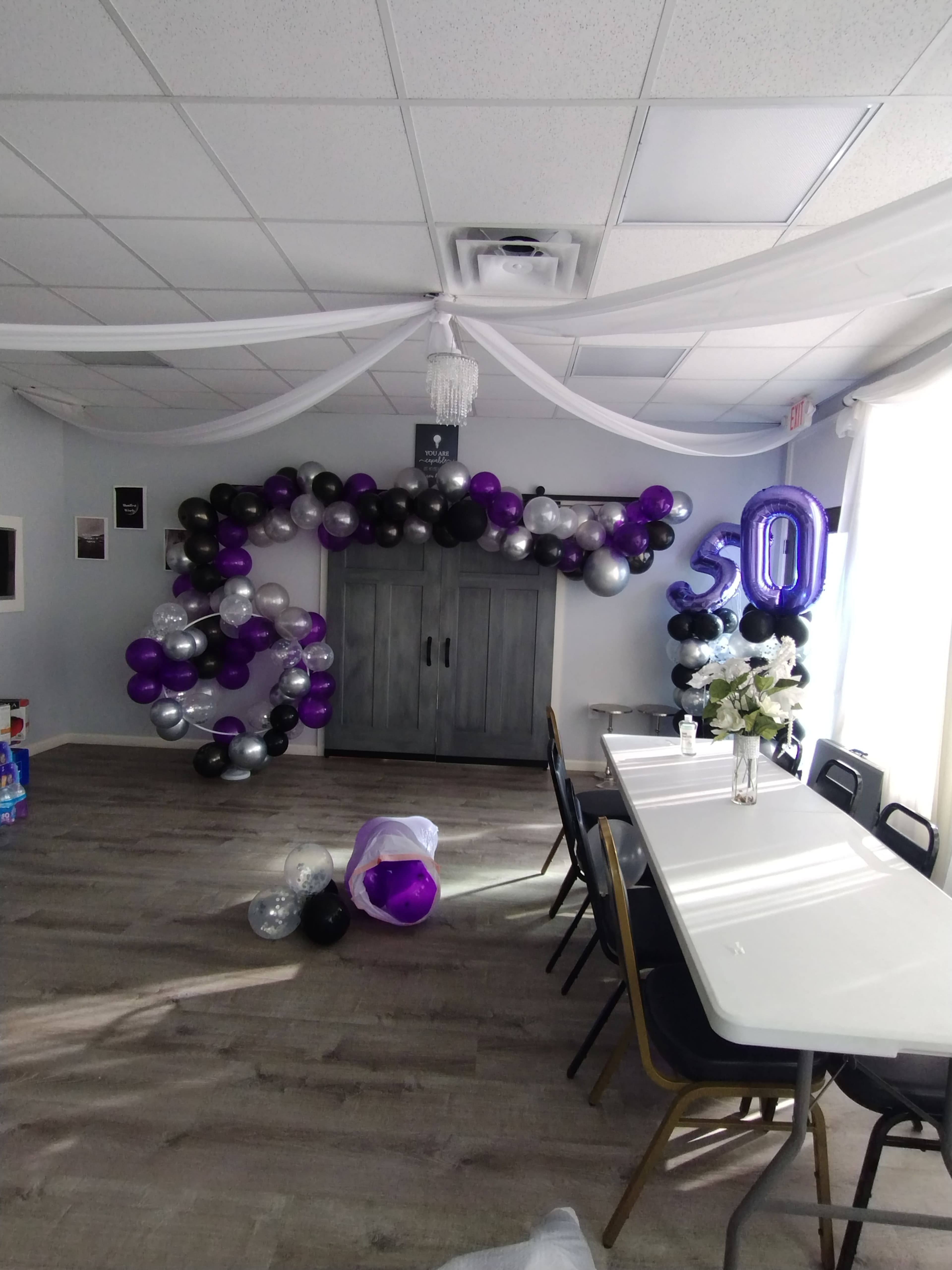 The room features a decorated wall with black, purple, and silver balloons arranged in an arch alongside a table set for an event, with a large "30" balloon displayed prominently.