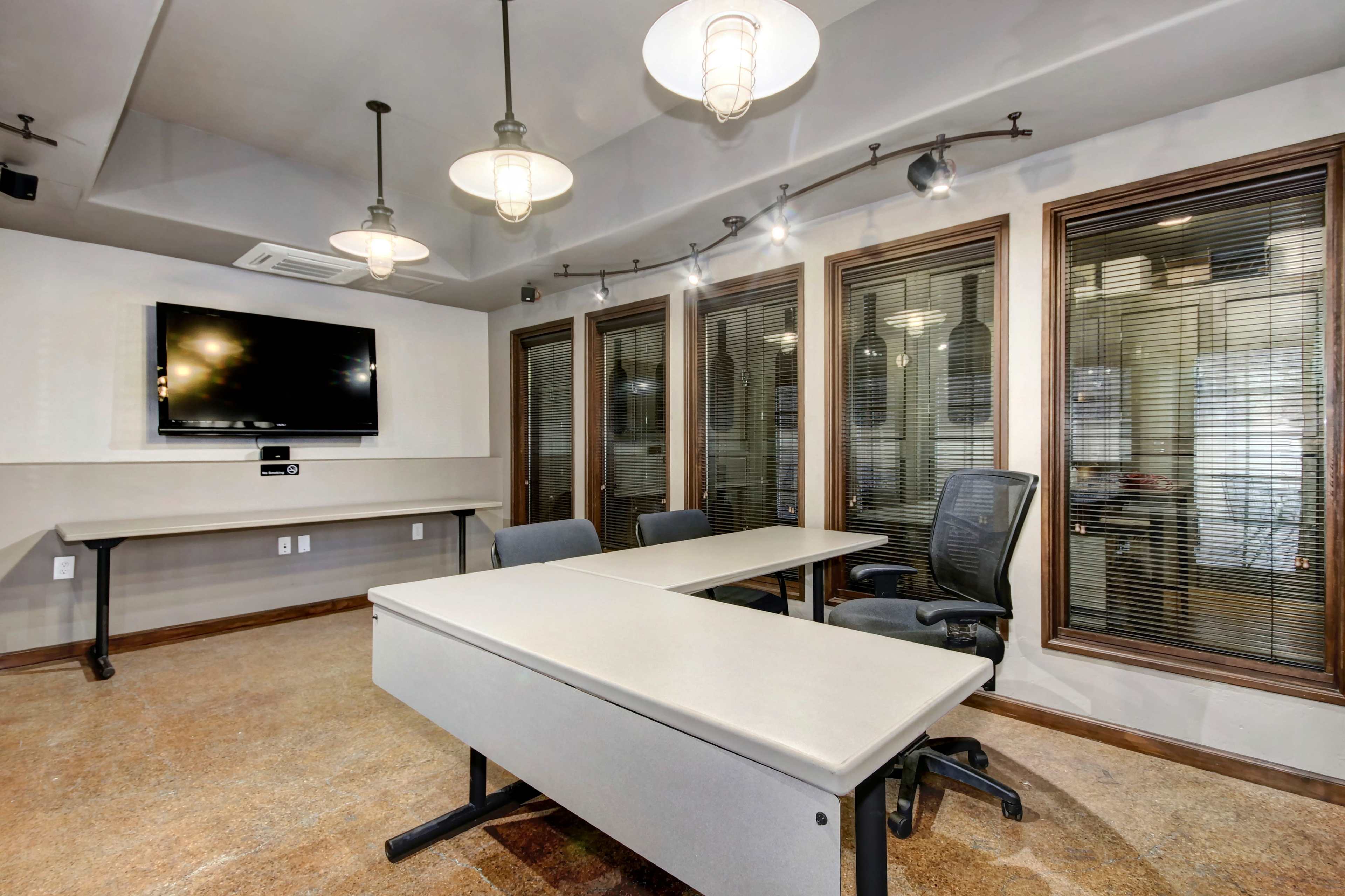 A modern conference room features a large table, ergonomic chairs, a wall-mounted television, and multiple windows with blinds.