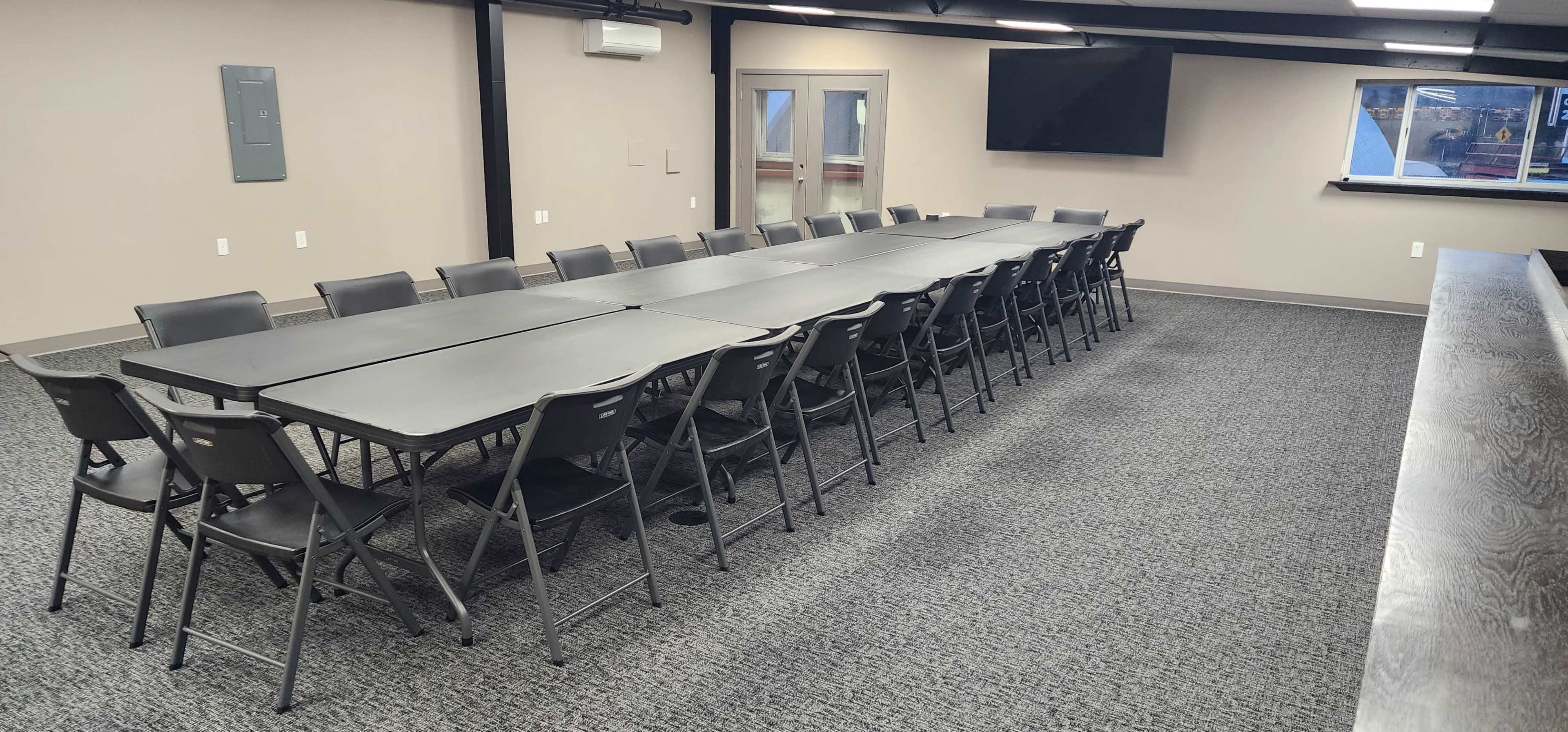 A long arrangement of folding tables and chairs fills a large, empty meeting room with a television and windows along one wall.