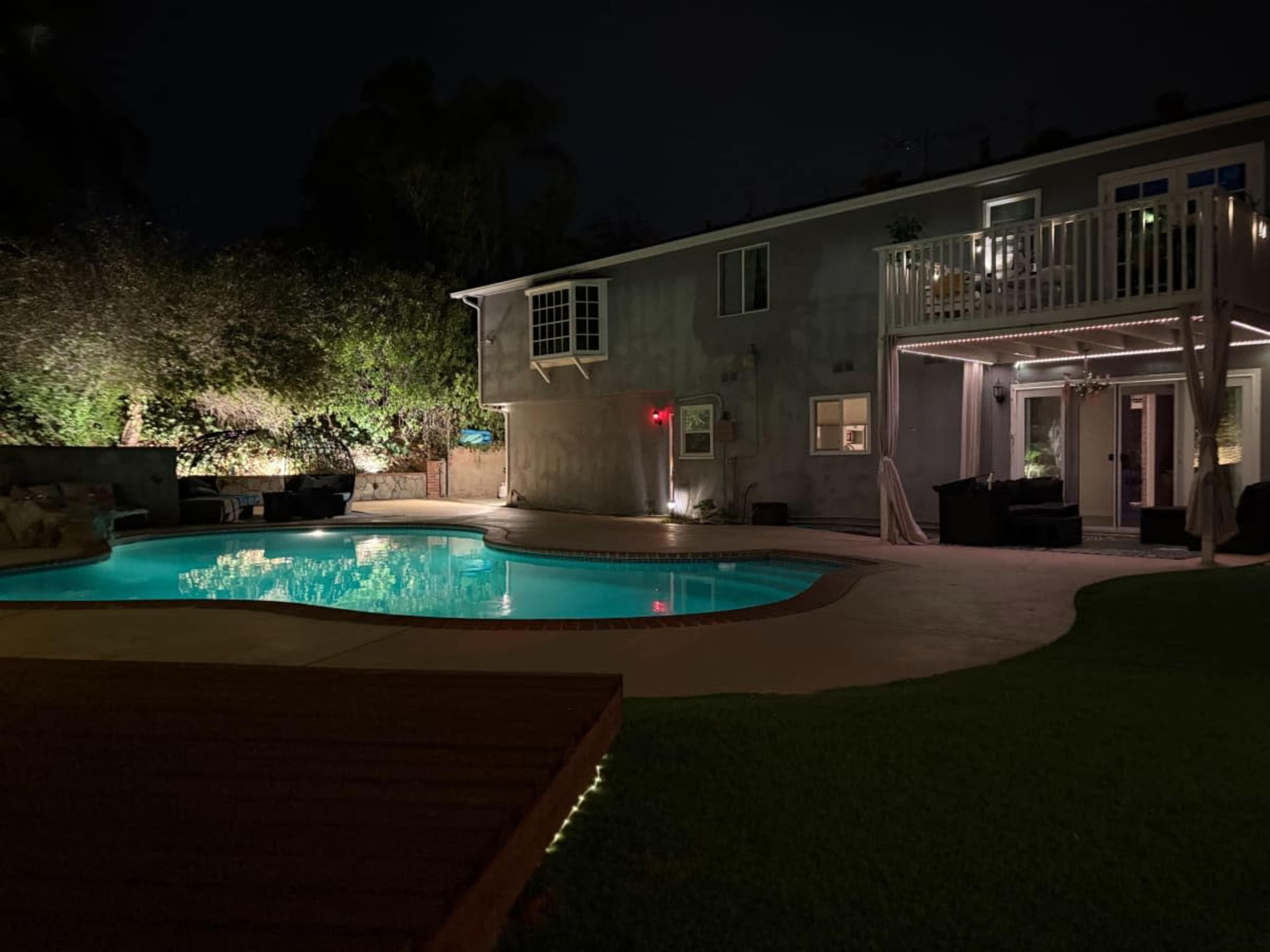 The image shows a well-lit backyard at night featuring a swimming pool in front of a two-story house with outdoor seating areas.