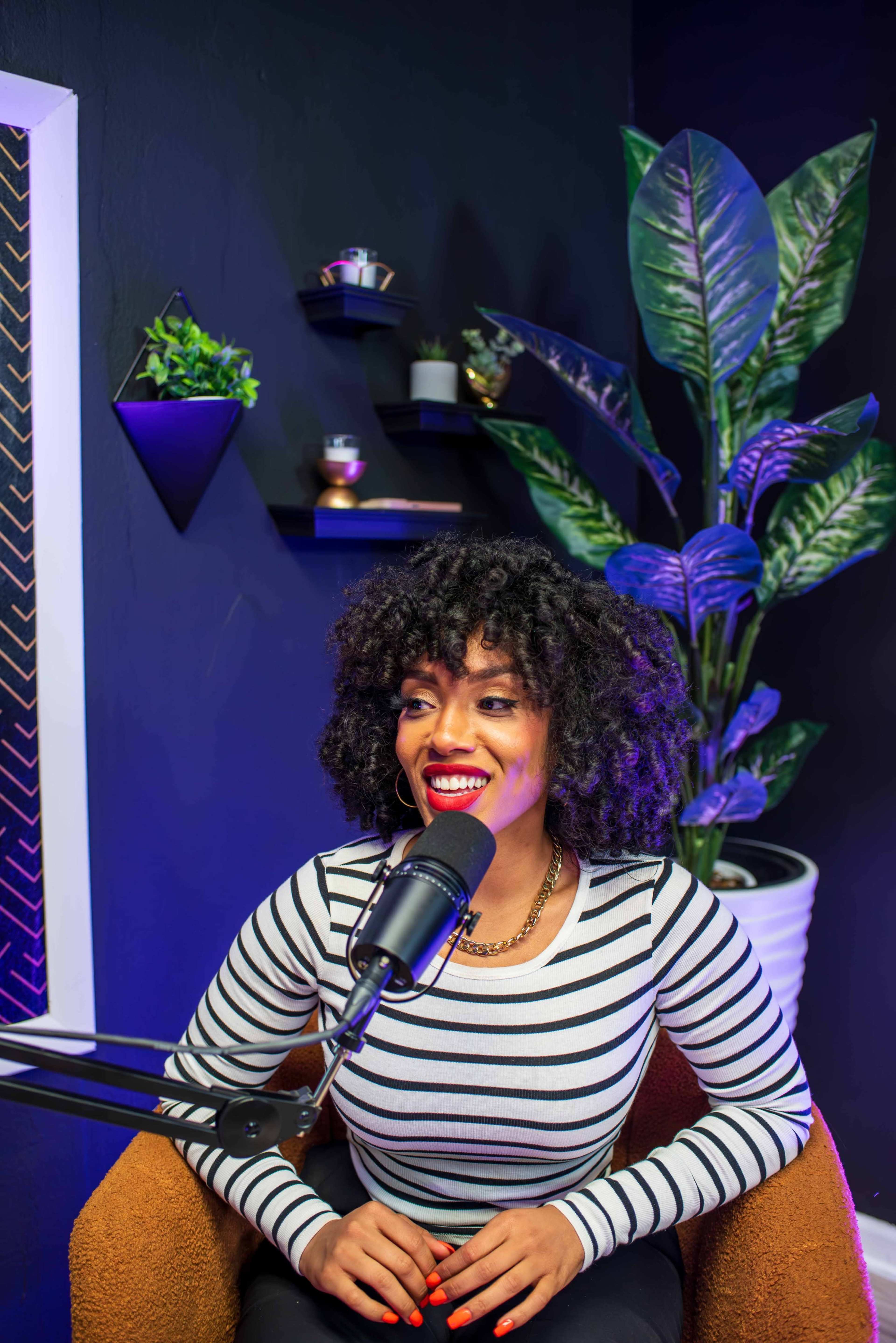 A woman with curly hair sits in front of a microphone in a studio decorated with plants and wall shelves.