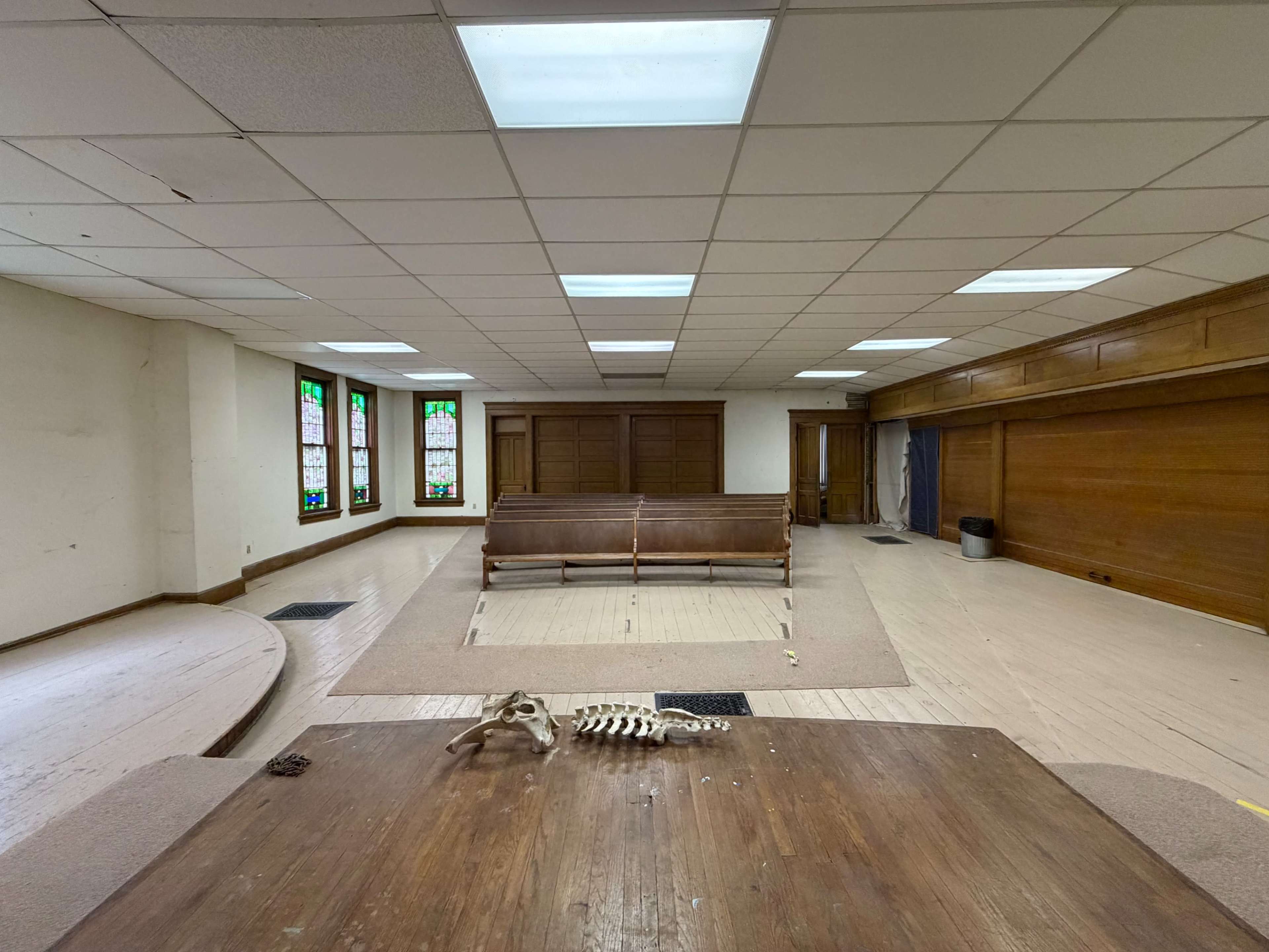 The image shows an empty room with wooden flooring, a large sectional area in the center, stained glass windows on the sides, and a small skeleton placed on a table.