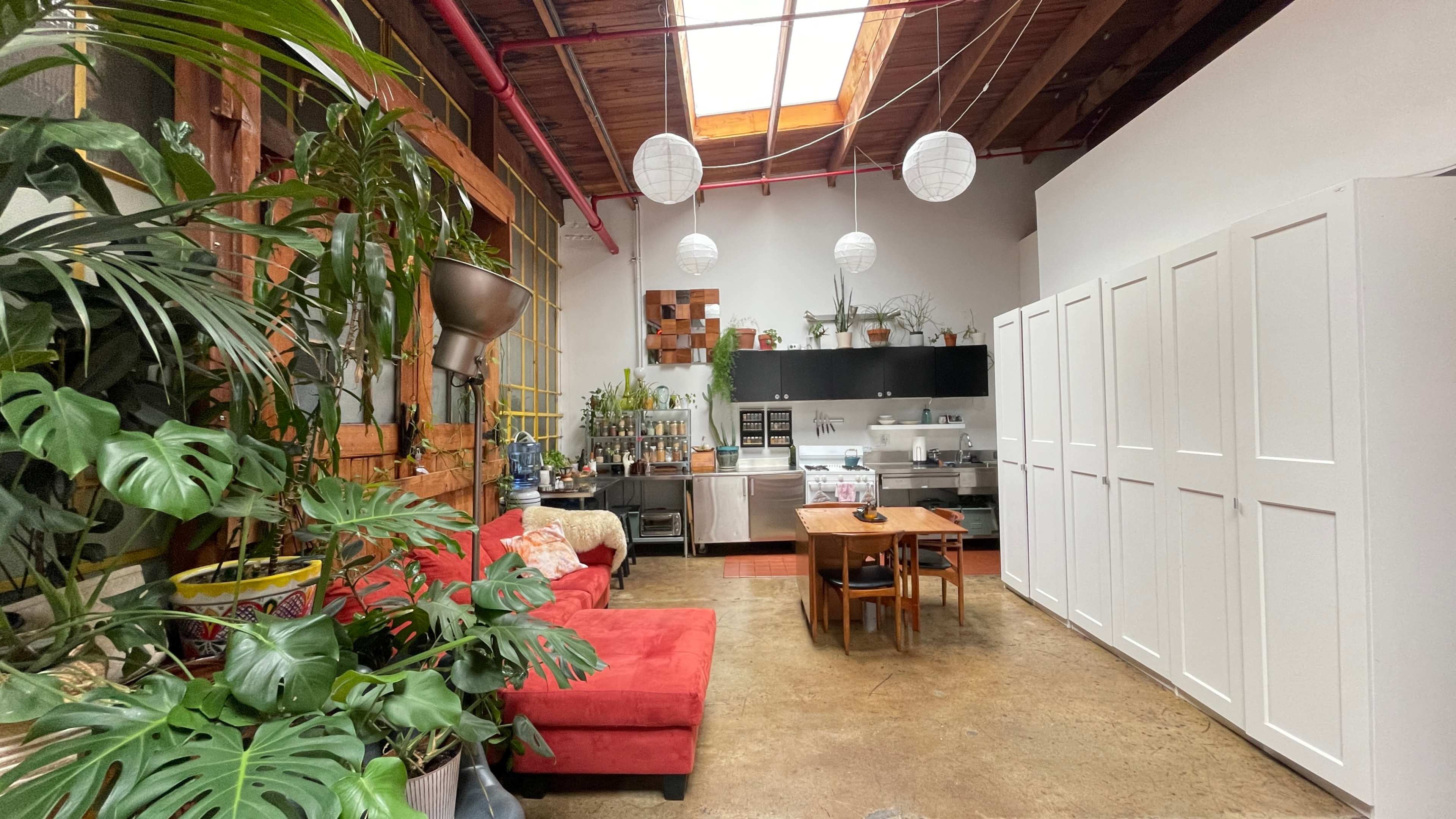 The image shows a spacious, modern kitchen and living area featuring a red sofa, a wooden dining table, and several indoor plants.