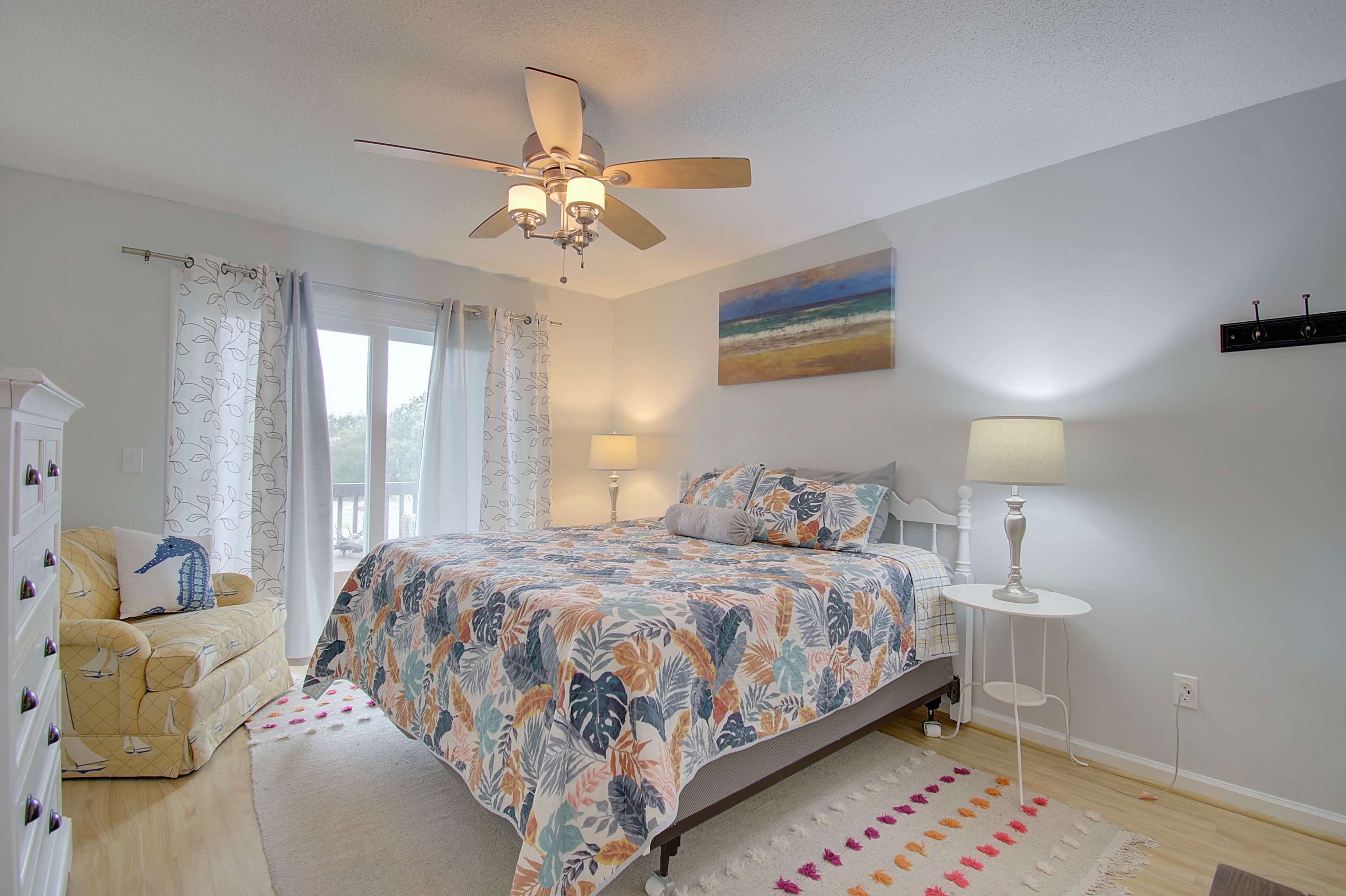 A bedroom with a large bed, a ceiling fan, two lamps, and a chair, all arranged against light-colored walls and a large window.