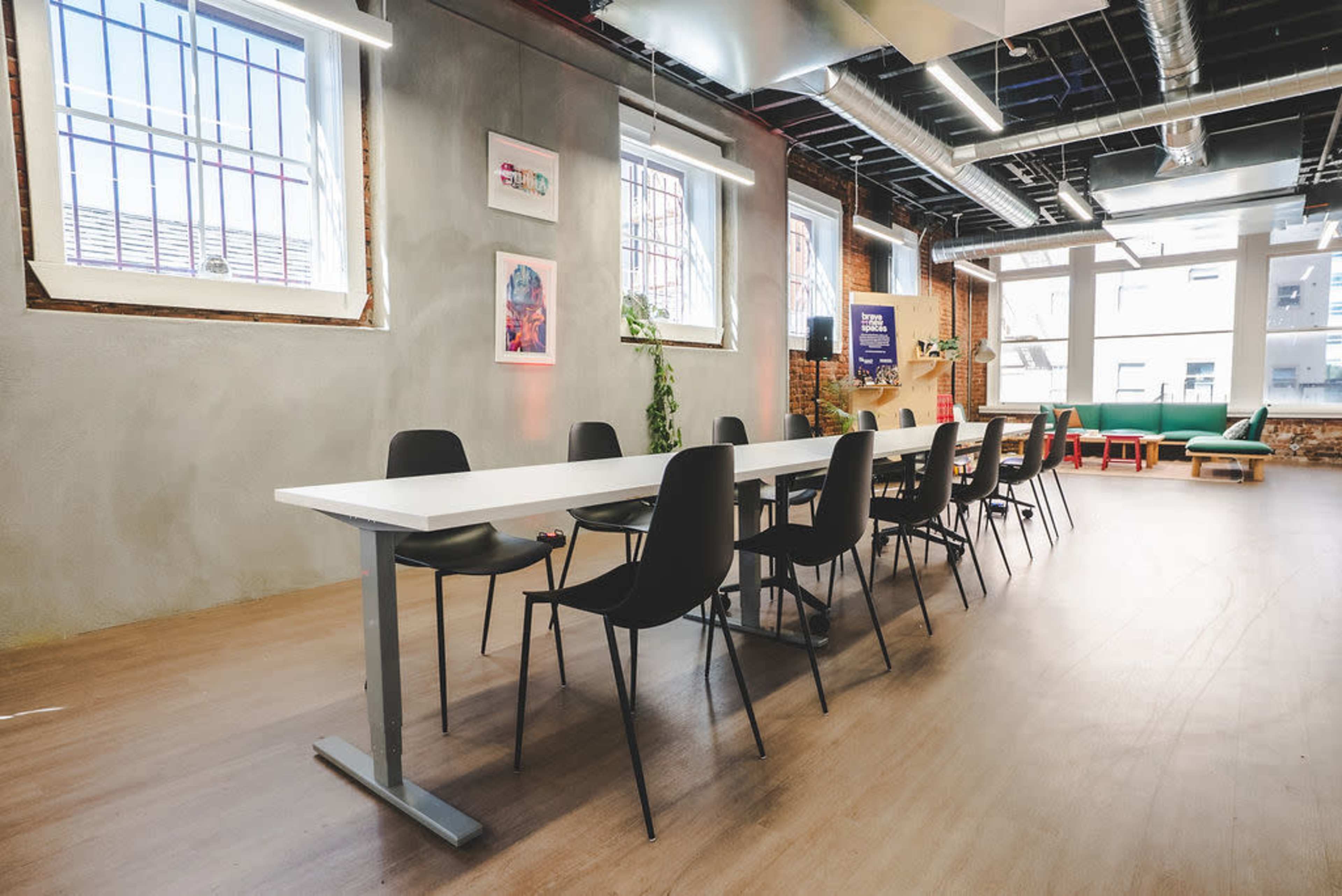 A long white table with black chairs is set up in a modern meeting room featuring exposed brick walls and large windows.