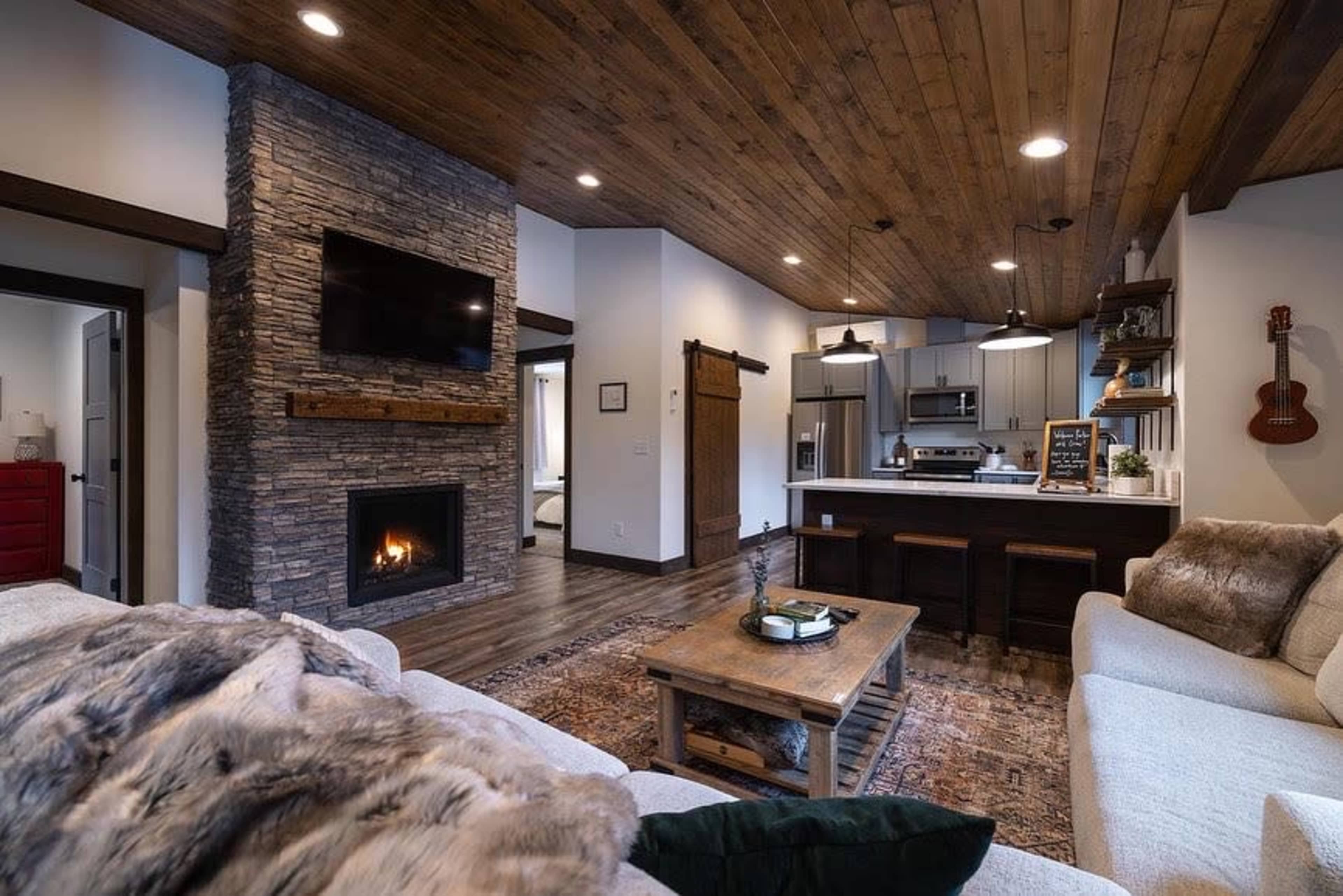 A modern living room with a stone fireplace, wooden ceiling beams, and a cozy seating area featuring a sofa and wooden coffee table.