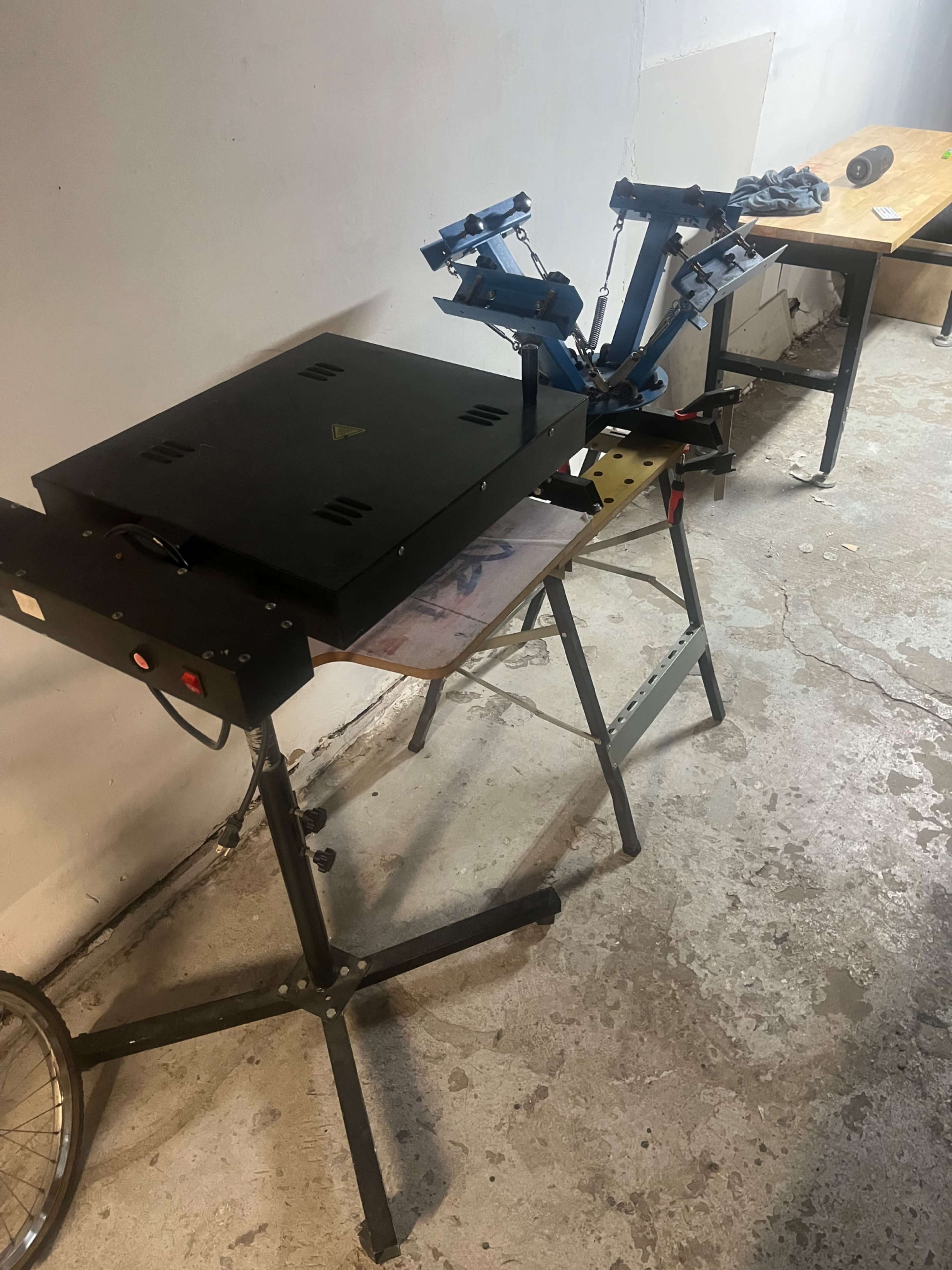 The image shows a workspace with a heat press machine and a screen printing setup placed on metal tables in a basement or industrial area.