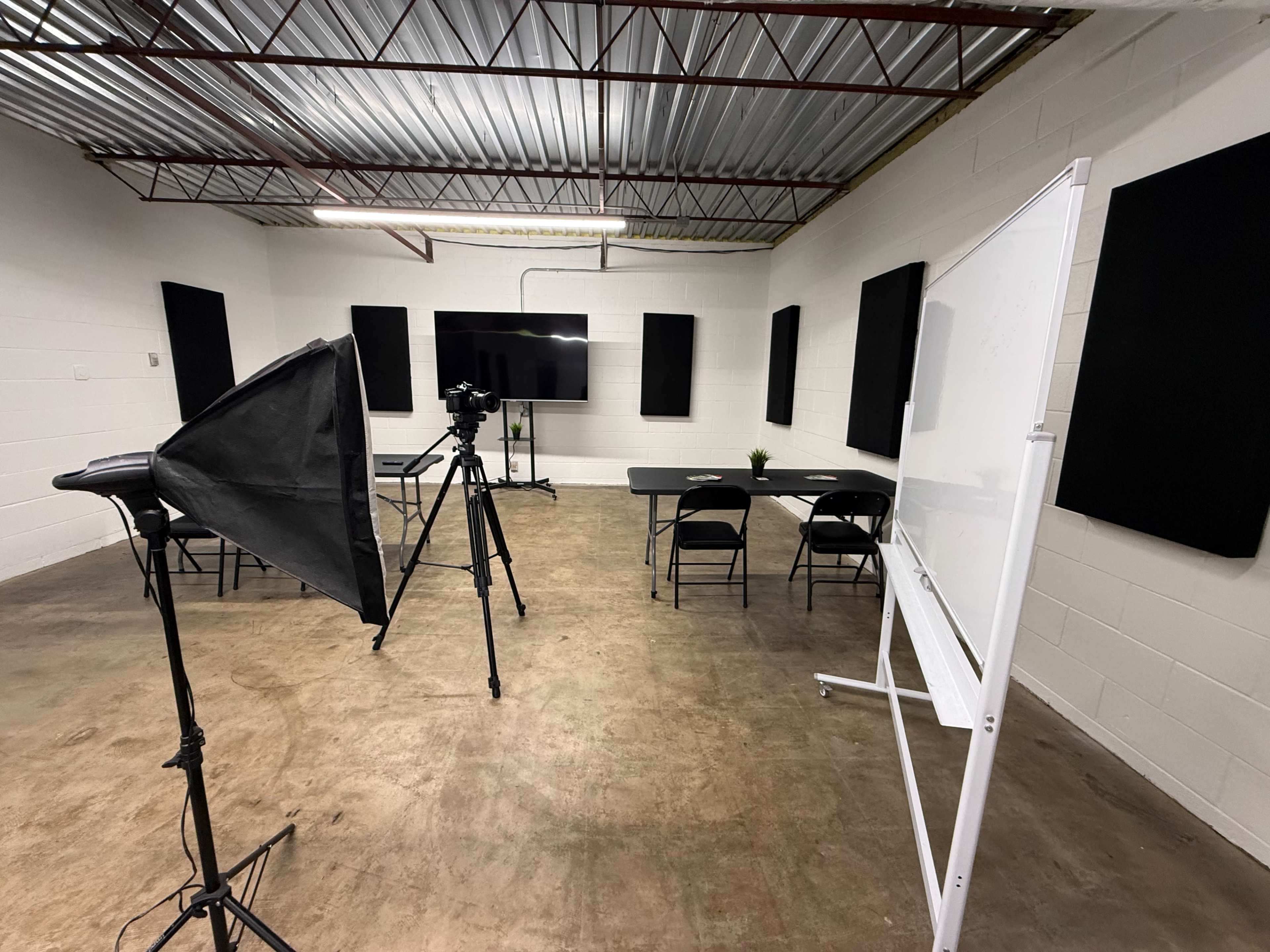 The image shows a minimalist room set up for a video recording, featuring a camera on a tripod, a large screen, a whiteboard, and several chairs arranged around a table.