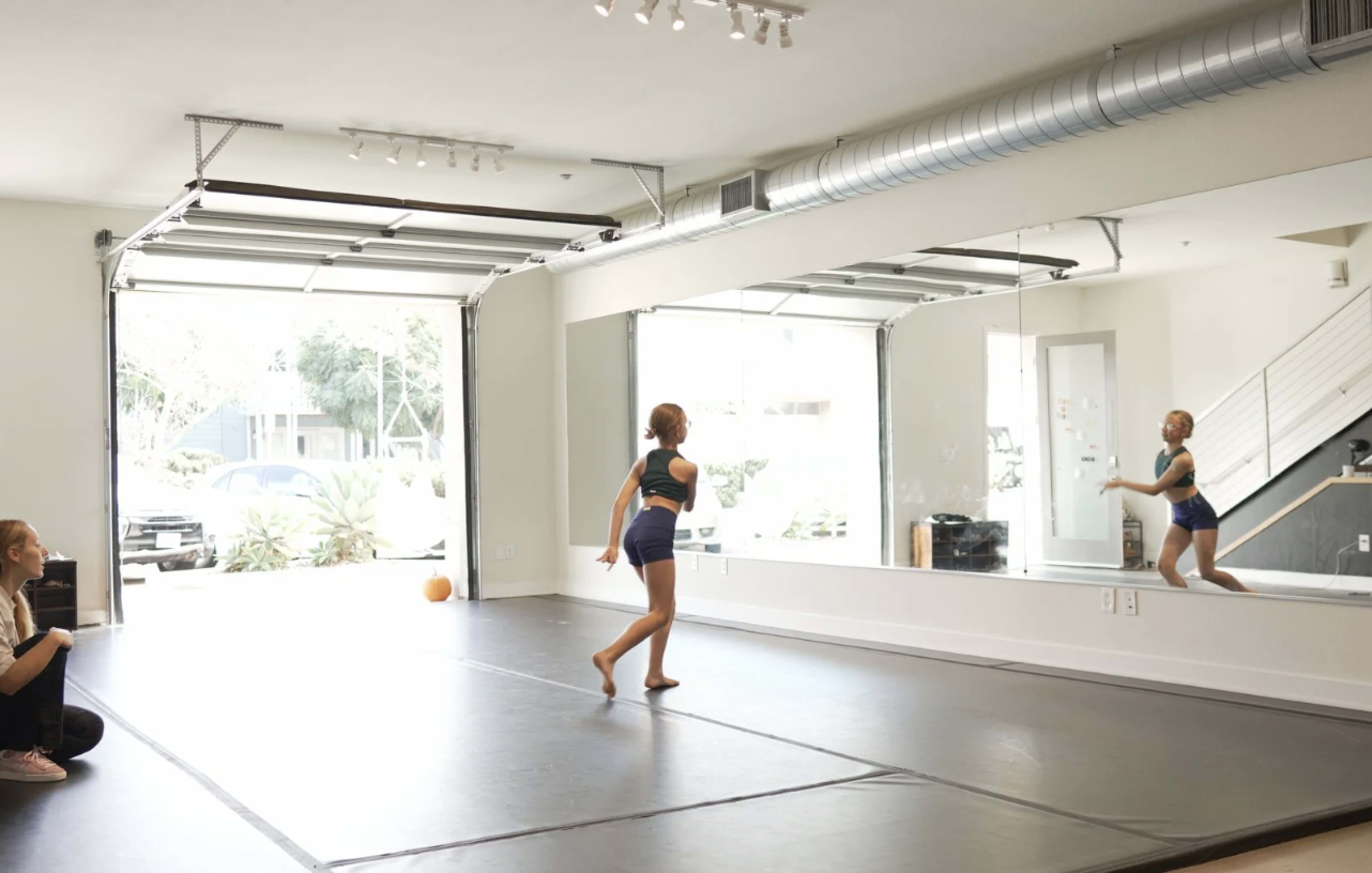 A dancer performs in a spacious studio with a large mirror, while an observer sits on the floor.